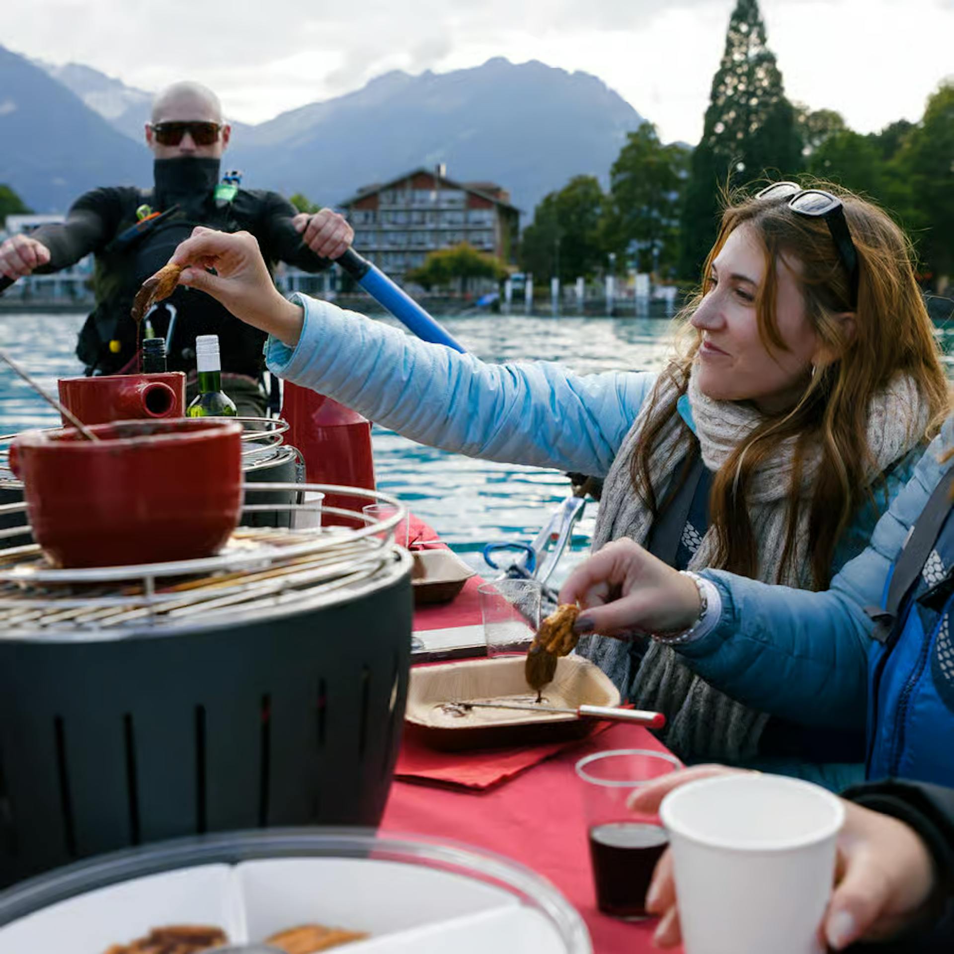 A woman dips a piece of food into a pot while enjoying a meal by the lakeside, surrounded by mountains and greenery.