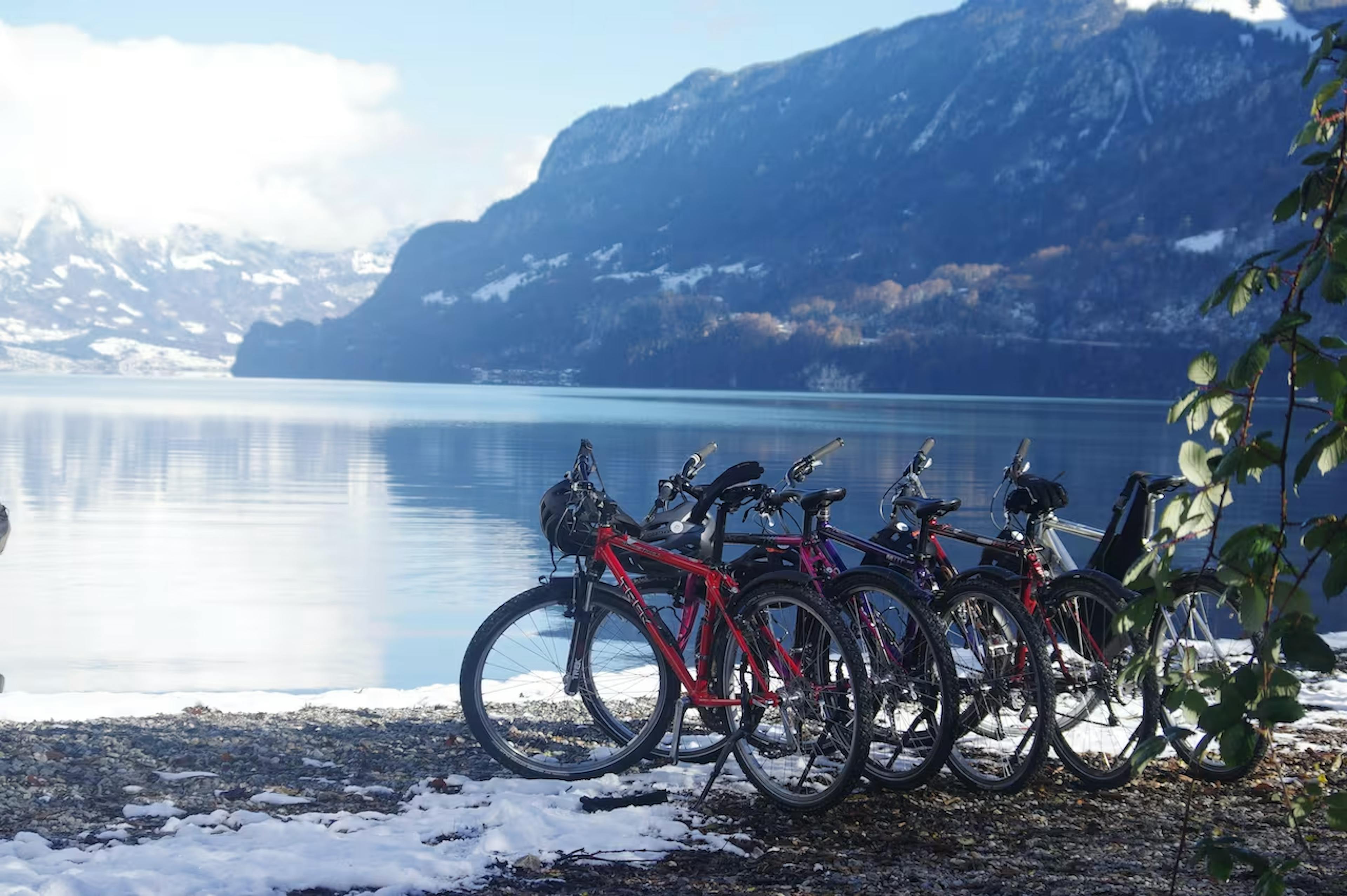 Bicycles are parked on a snowy lakeside shore with a backdrop of placid water and snow-capped mountains under a clear blue sky. Serene and scenic.