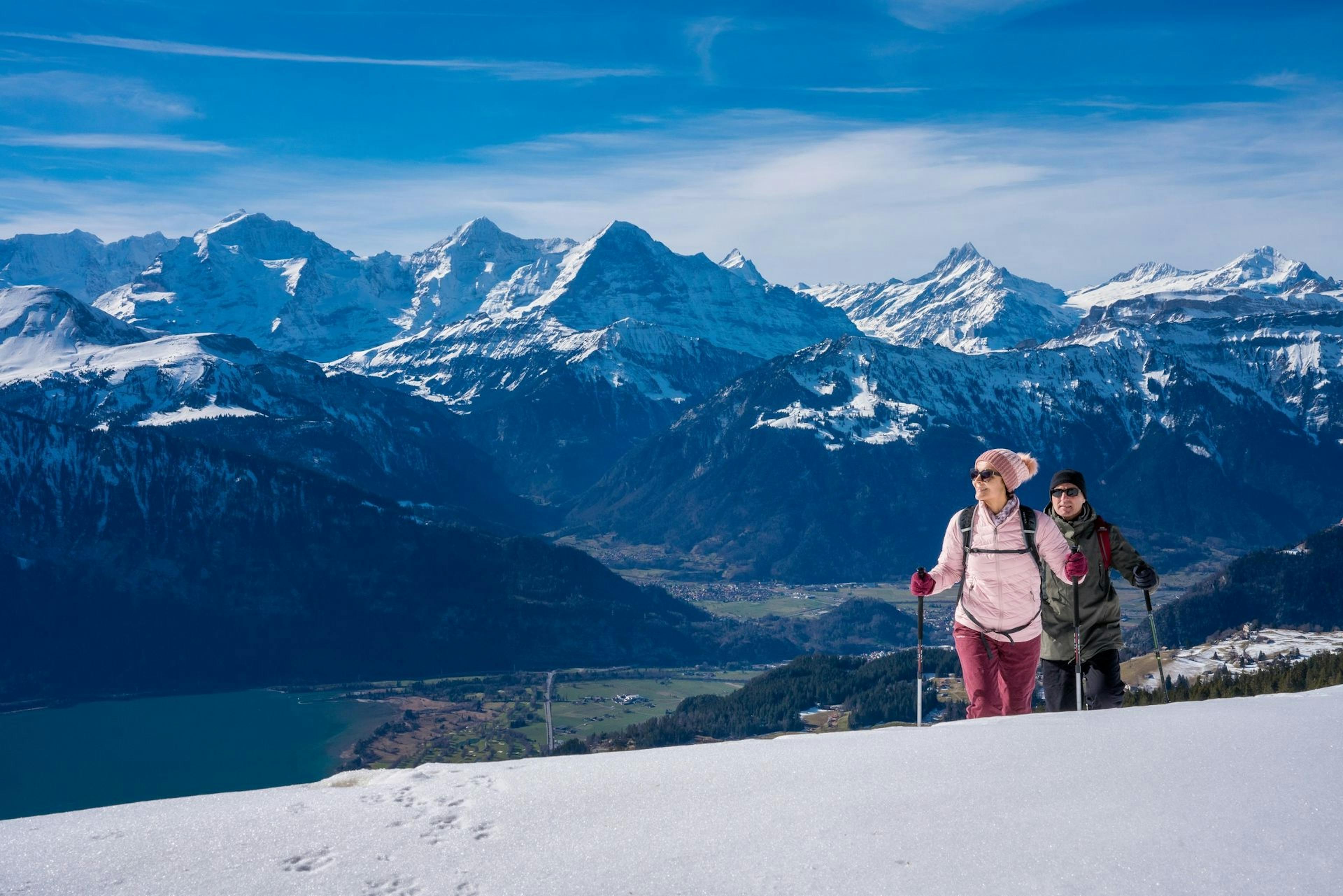 Two people hike uphill on a snowy slope with a scenic backdrop of snow-capped mountains, blue sky, and a lake below.