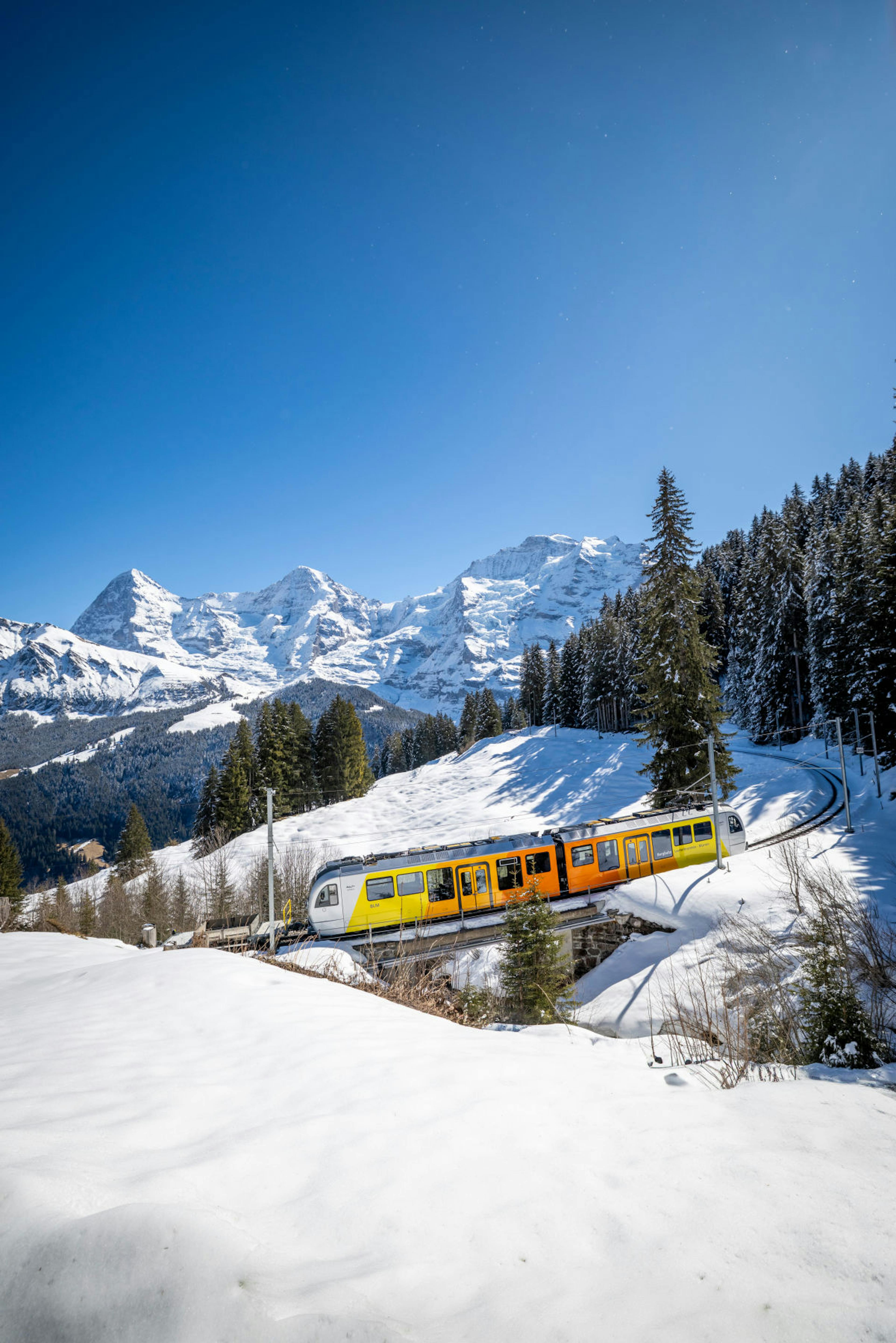 A bright yellow and orange train travels through a snowy landscape. Snow-covered mountains and evergreen trees are set against a clear blue sky. Peaceful winter scene.
