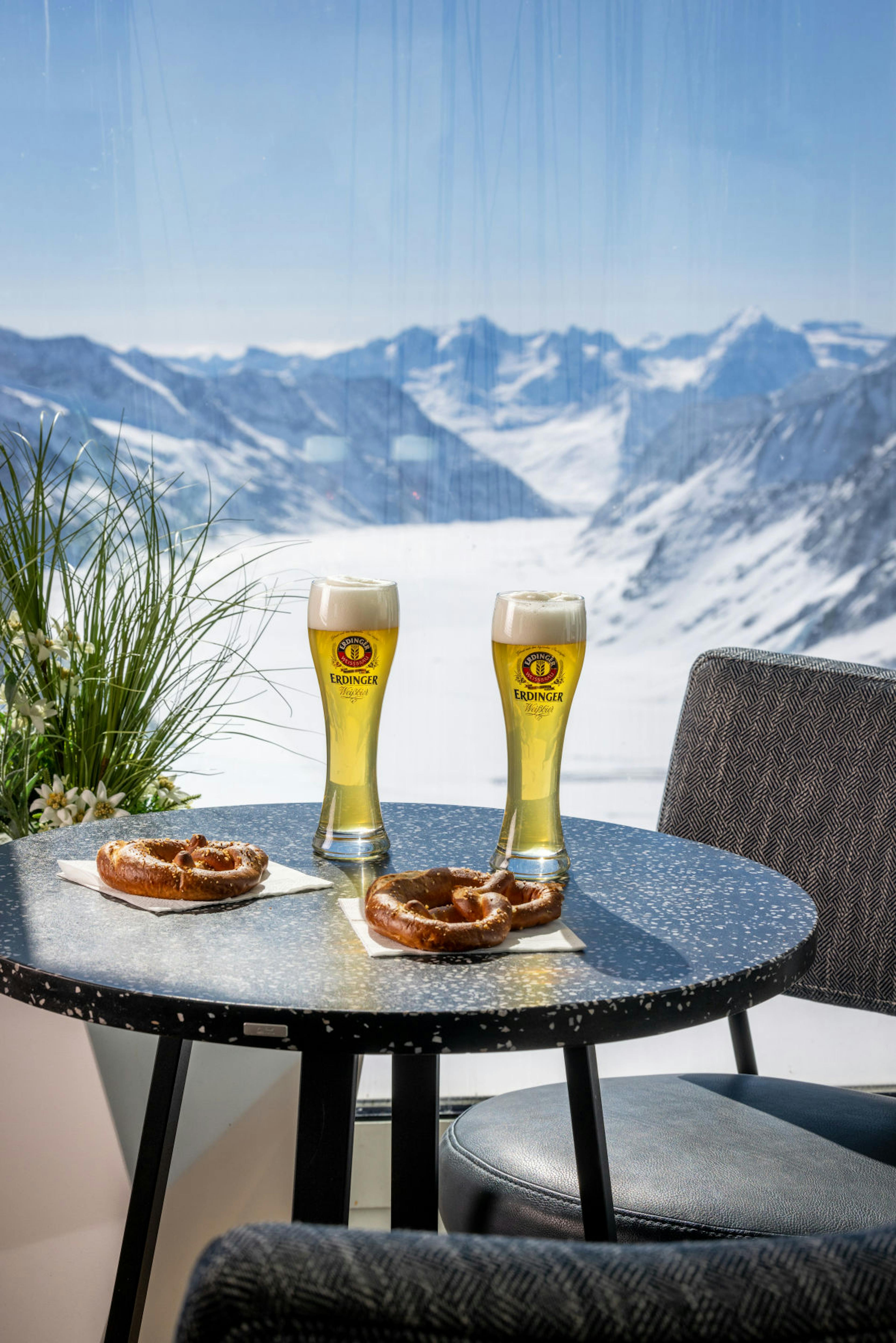 Two tall glasses of beer and pretzels sit on a table with a snowy mountain backdrop. Bright, clear sky adds a fresh, serene atmosphere.