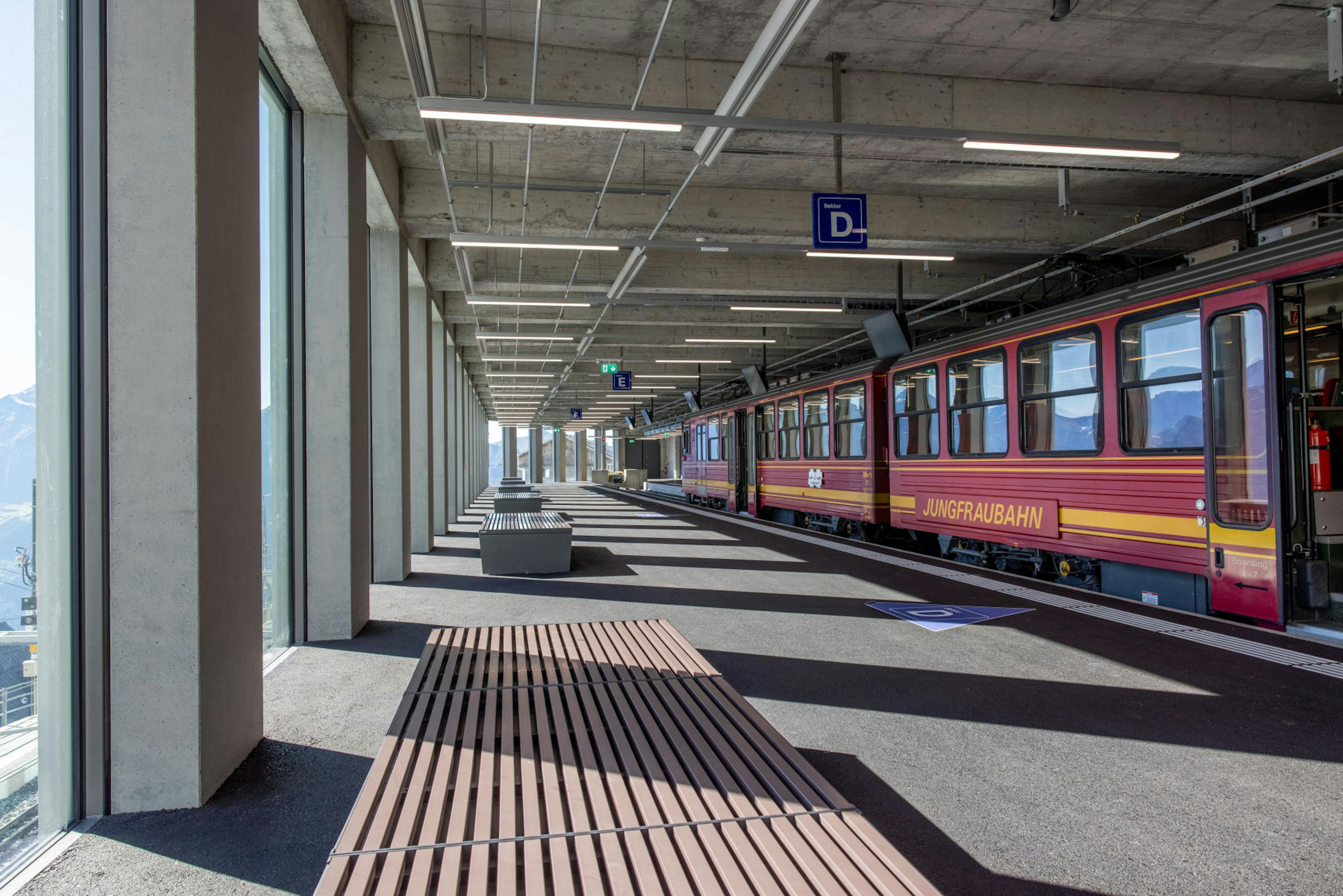 Train station platform with a red Jungfraubahn train on the right. Sunlit modern architecture with large windows and wooden benches. Calm atmosphere.