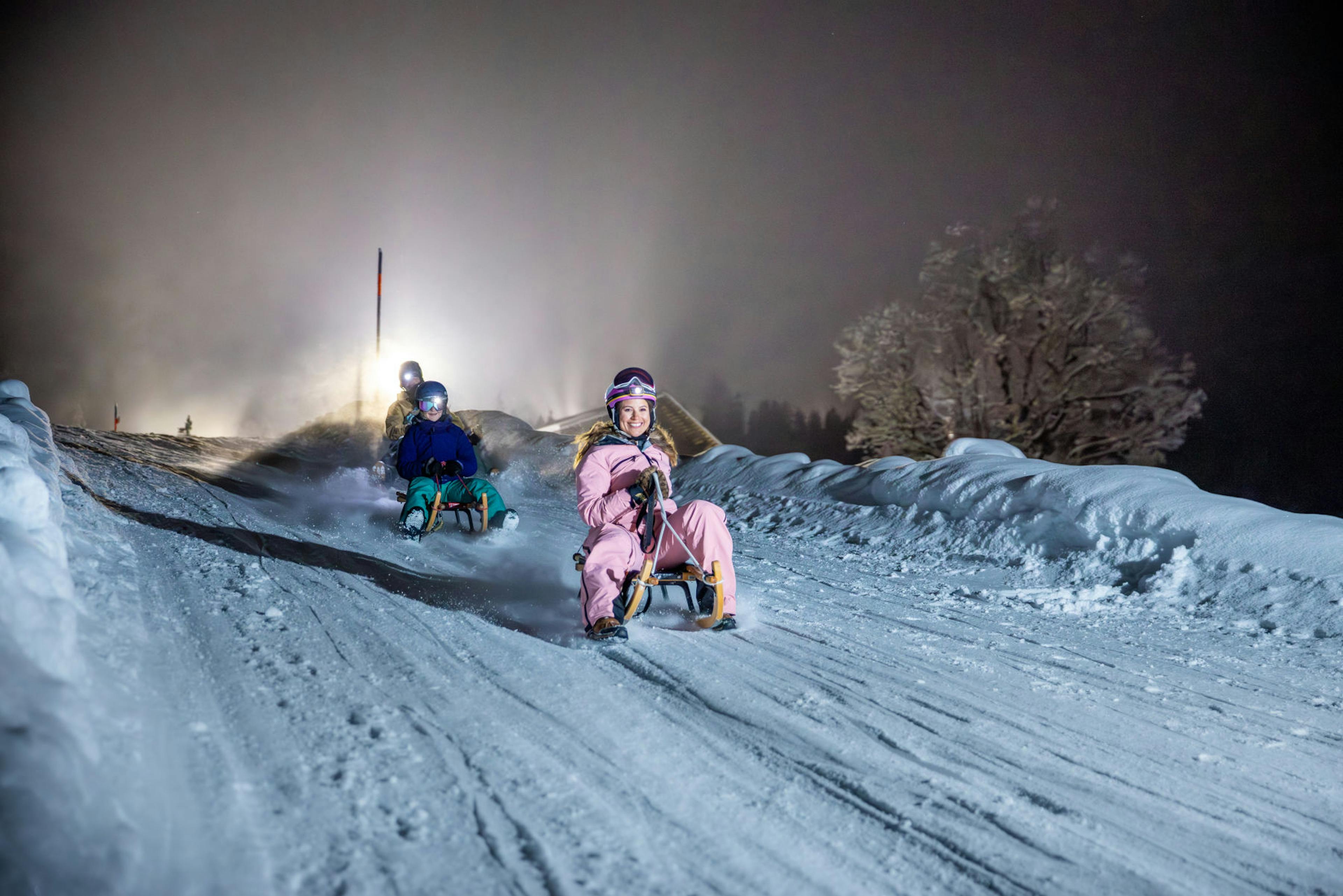 Children sled down a snowy hill at night, illuminated by a bright light behind them. The scene conveys excitement and winter fun, with snow-covered trees to the right.