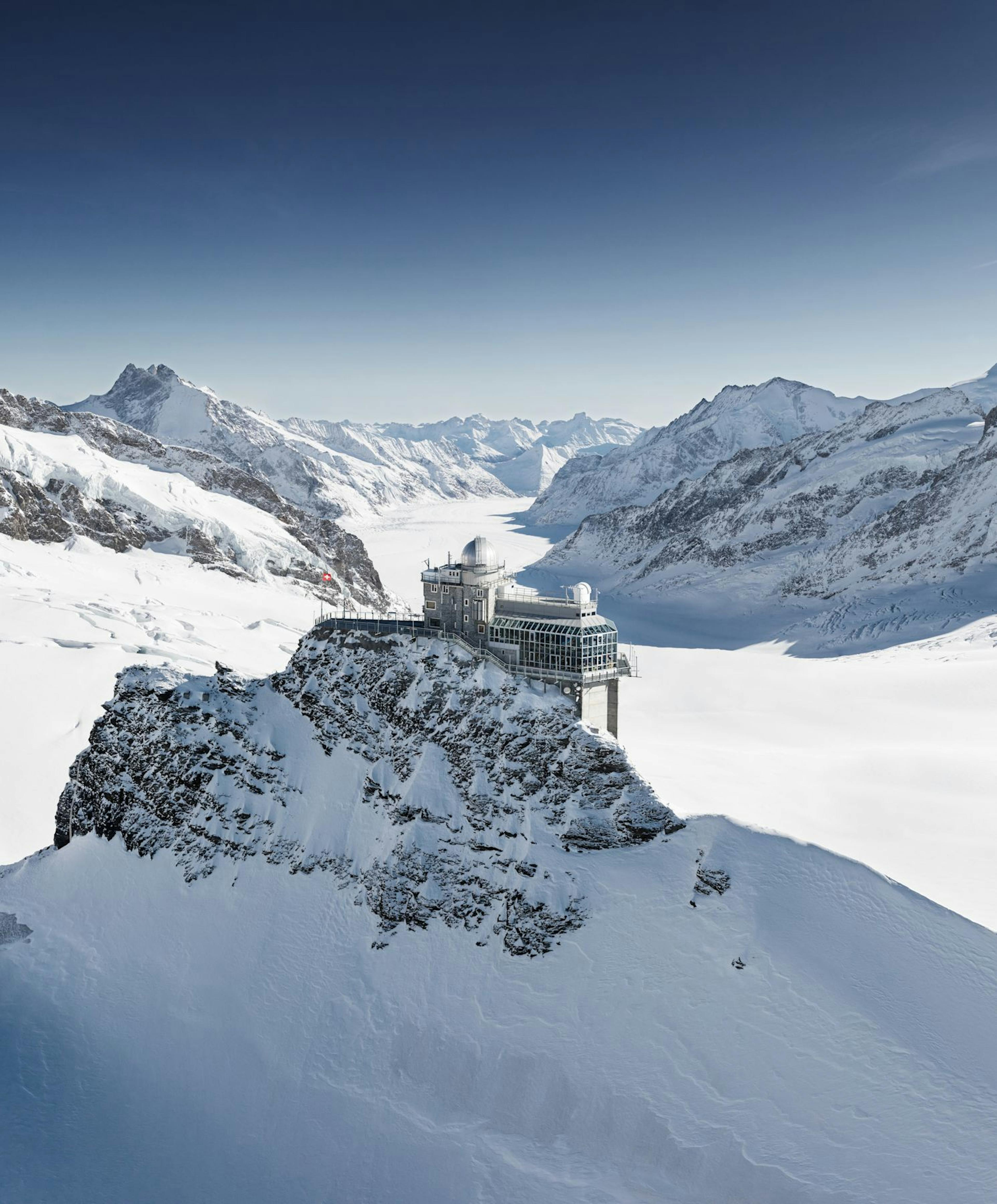 Snowy mountain landscape with a modern observatory perched on a peak, surrounded by vast white slopes under a clear blue sky. Majestic and serene atmosphere.