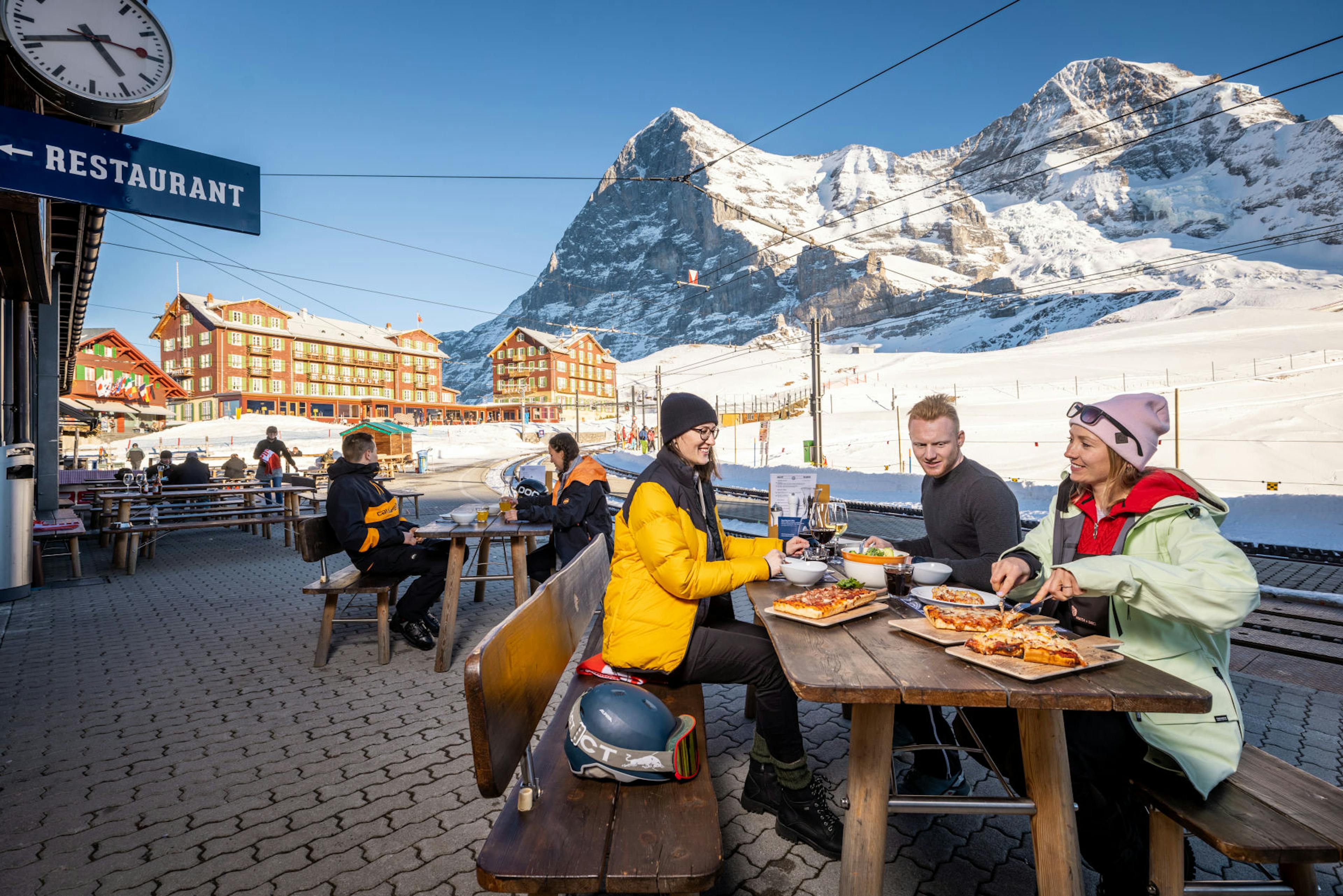 People enjoying a meal at an outdoor restaurant in a snowy mountain setting. Bright, sunny day with clear blue skies, creating a cheerful atmosphere.