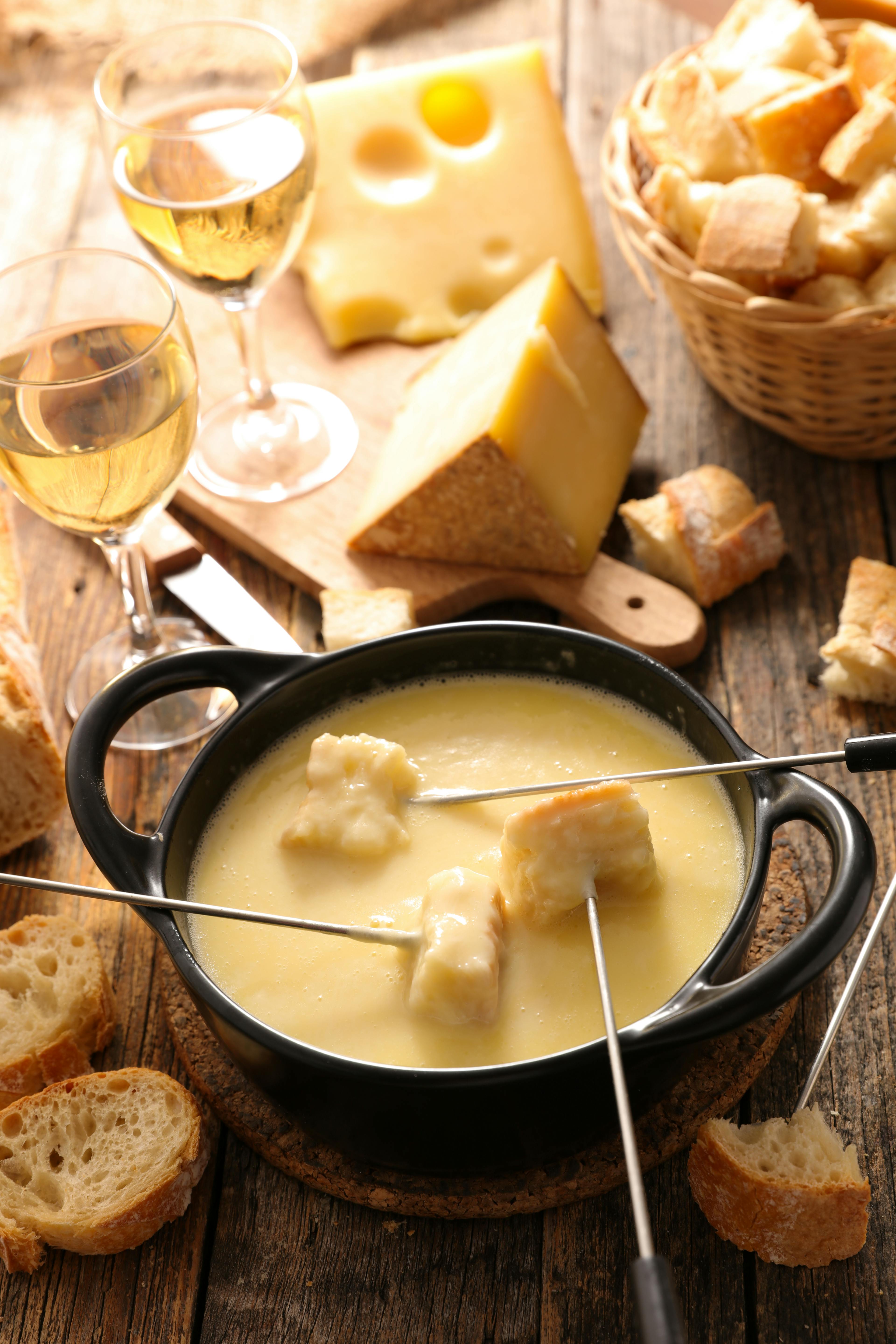 Cheese fondue with bread cubes on skewers in a black pot, surrounded by sliced bread, cheese blocks, and two glasses of white wine on a rustic table.