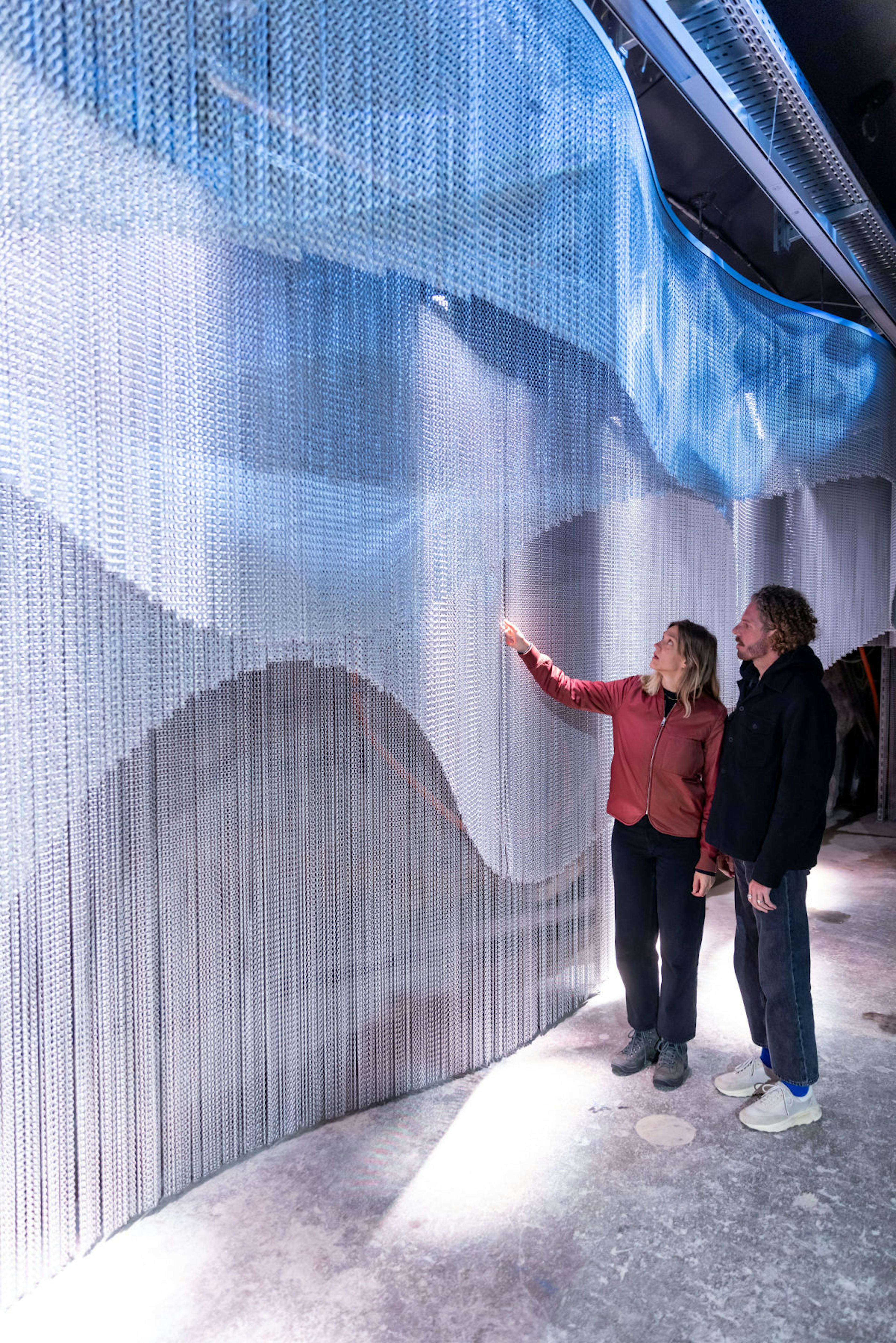 Two people admire a large, wavy installation made of hanging chains with blue lighting. The atmosphere is curious and contemplative.