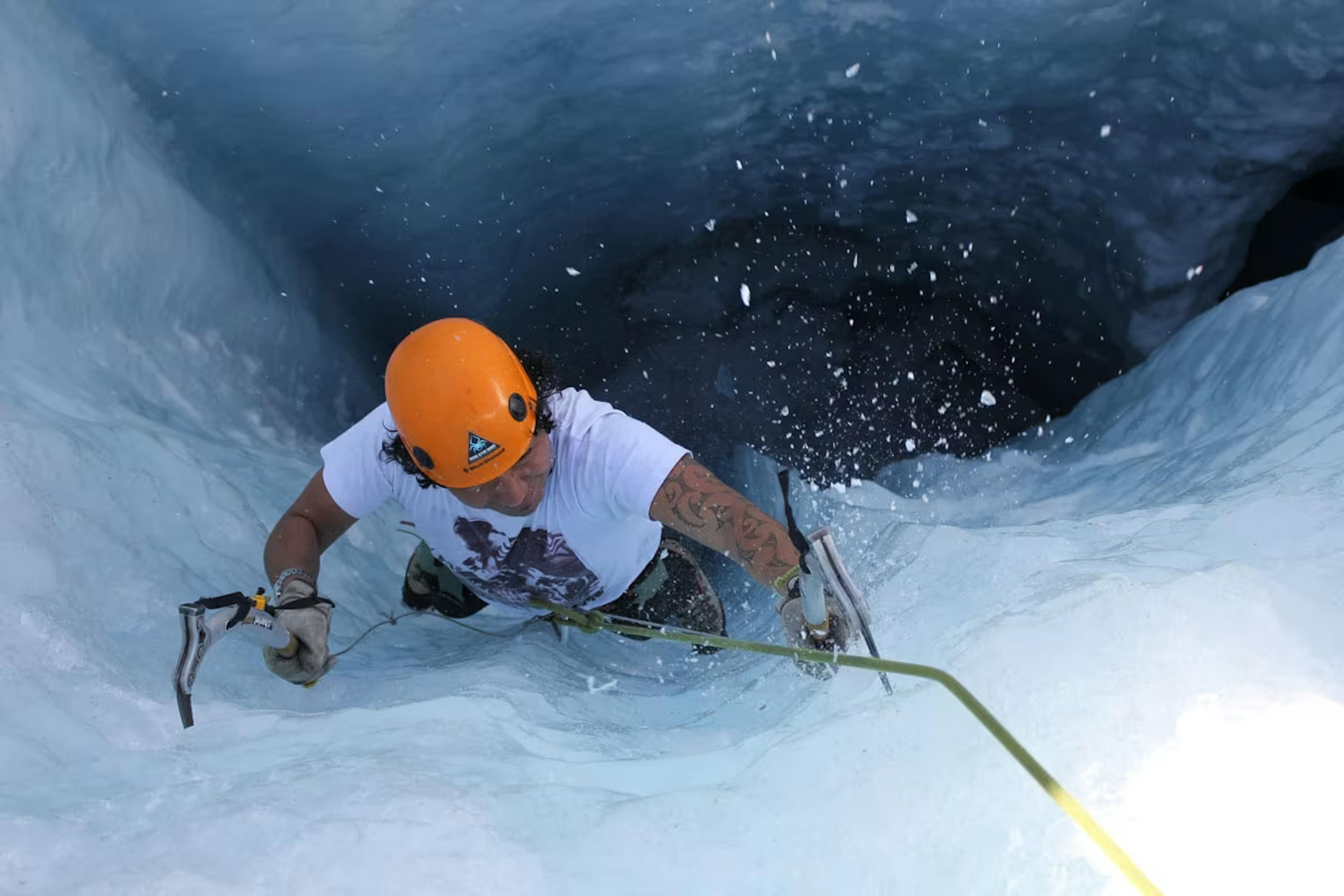 A climber with an orange helmet and ice axes ascends a steep, icy slope. Snowflakes scatter around, conveying a sense of challenge and adventure.