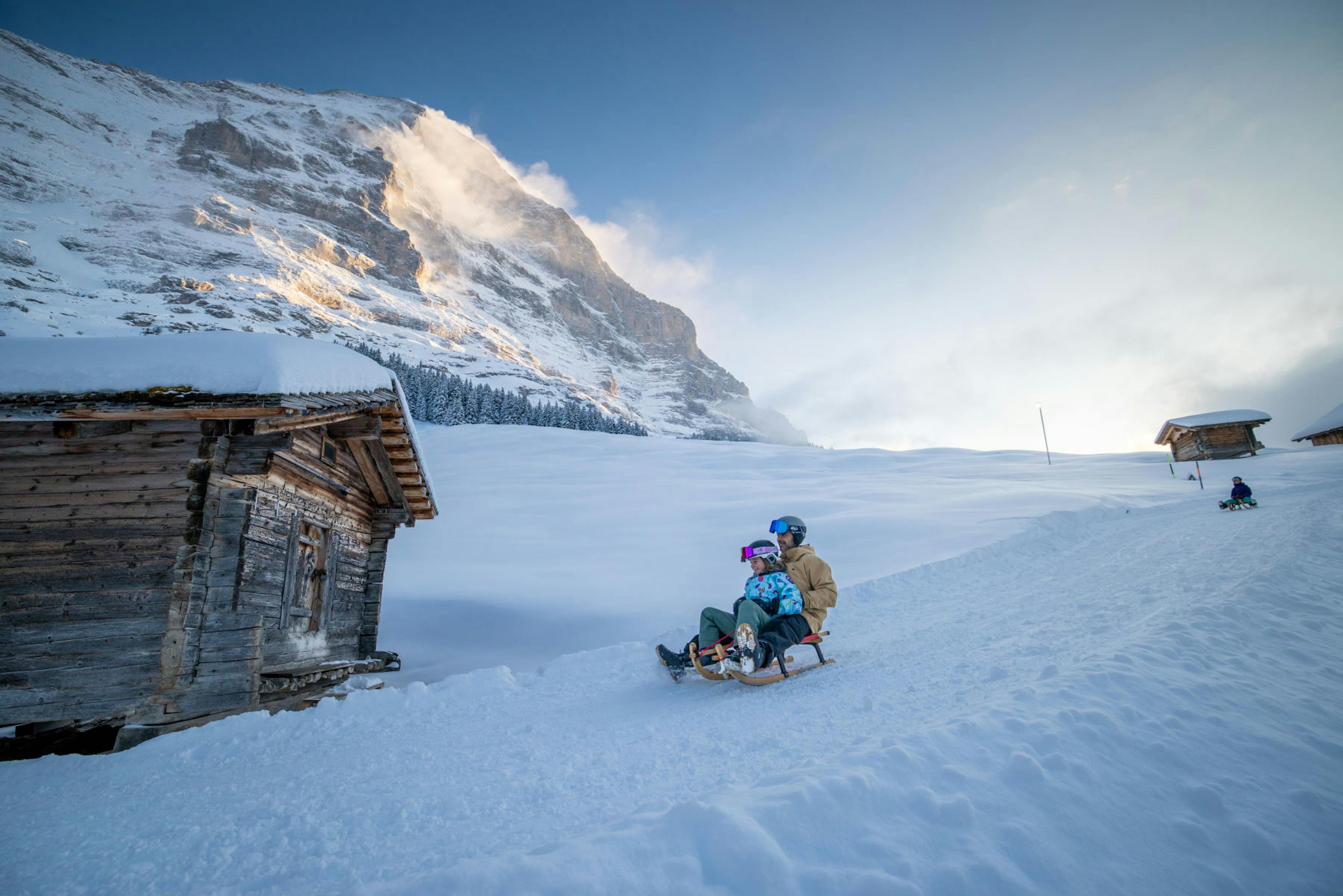 People sledding down a snowy hill in a scenic alpine landscape. Two cabins appear left and right. Majestic mountain under a clear blue sky.