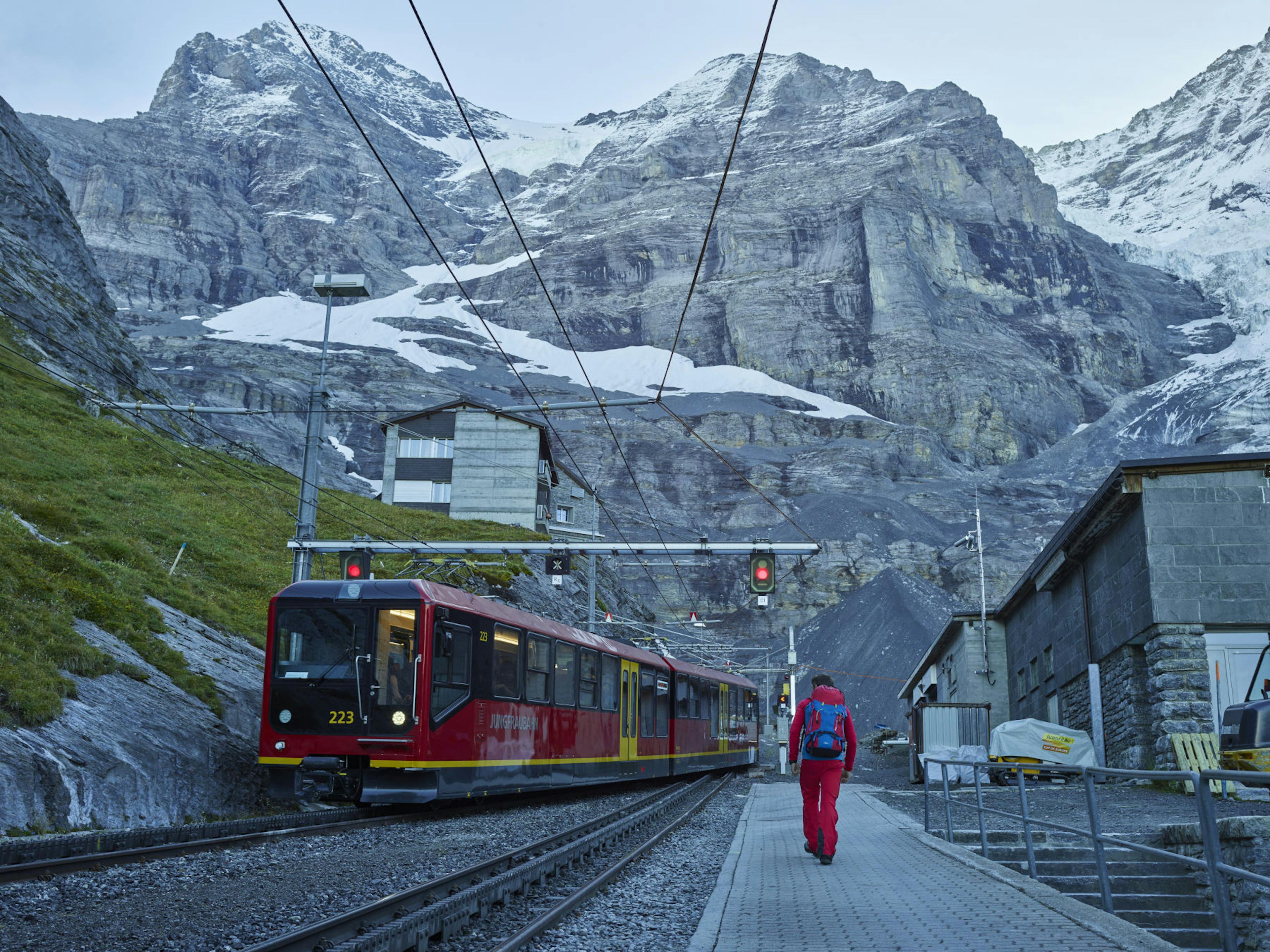 Red train on mountain tracks with a backdrop of rugged snow-dusted peaks. A person in a red outfit walks along the platform, conveying a sense of adventure.