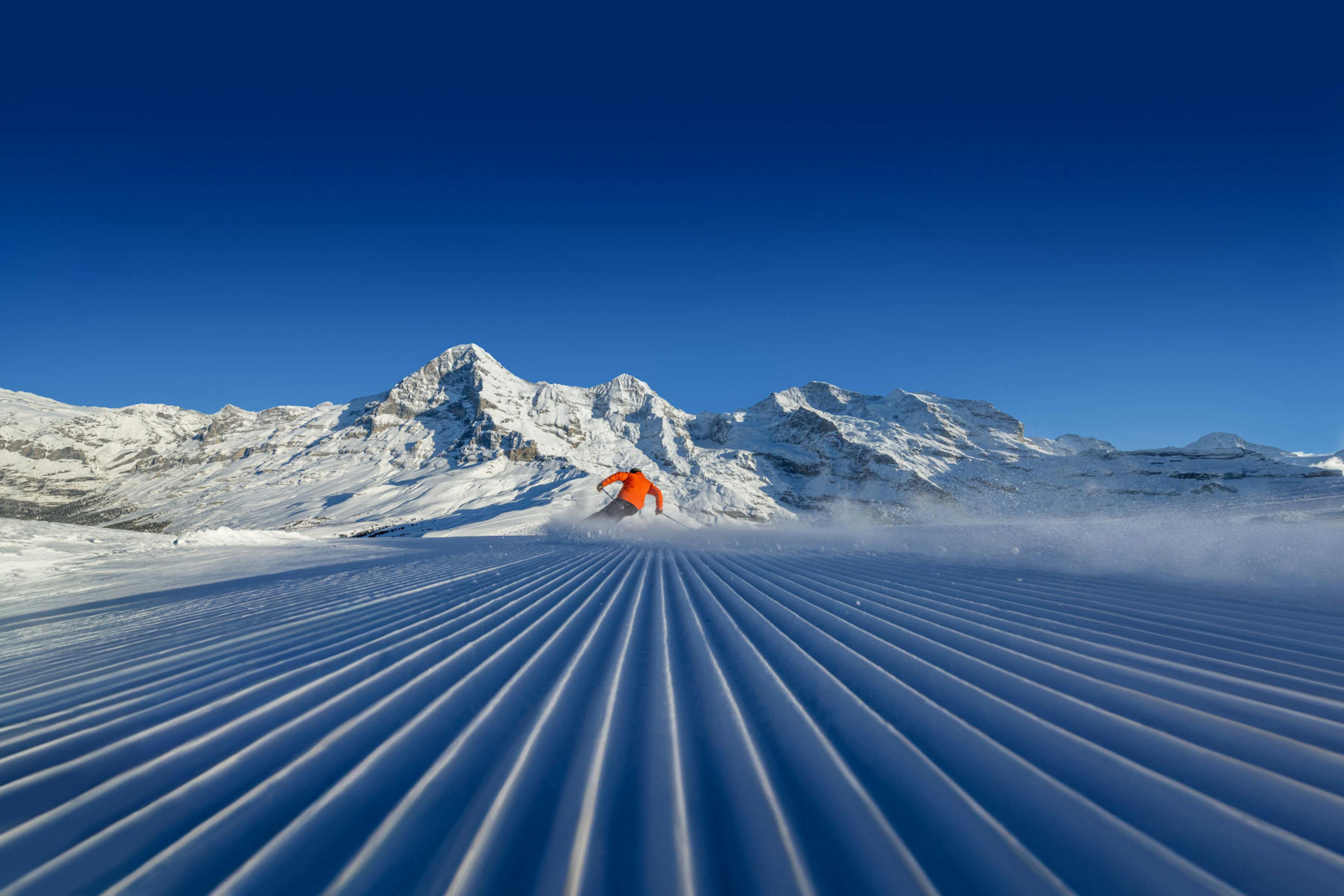 Skier in orange jacket swiftly descends a groomed snowy slope with distant snow-covered mountains under a clear blue sky, conveying speed and exhilaration.