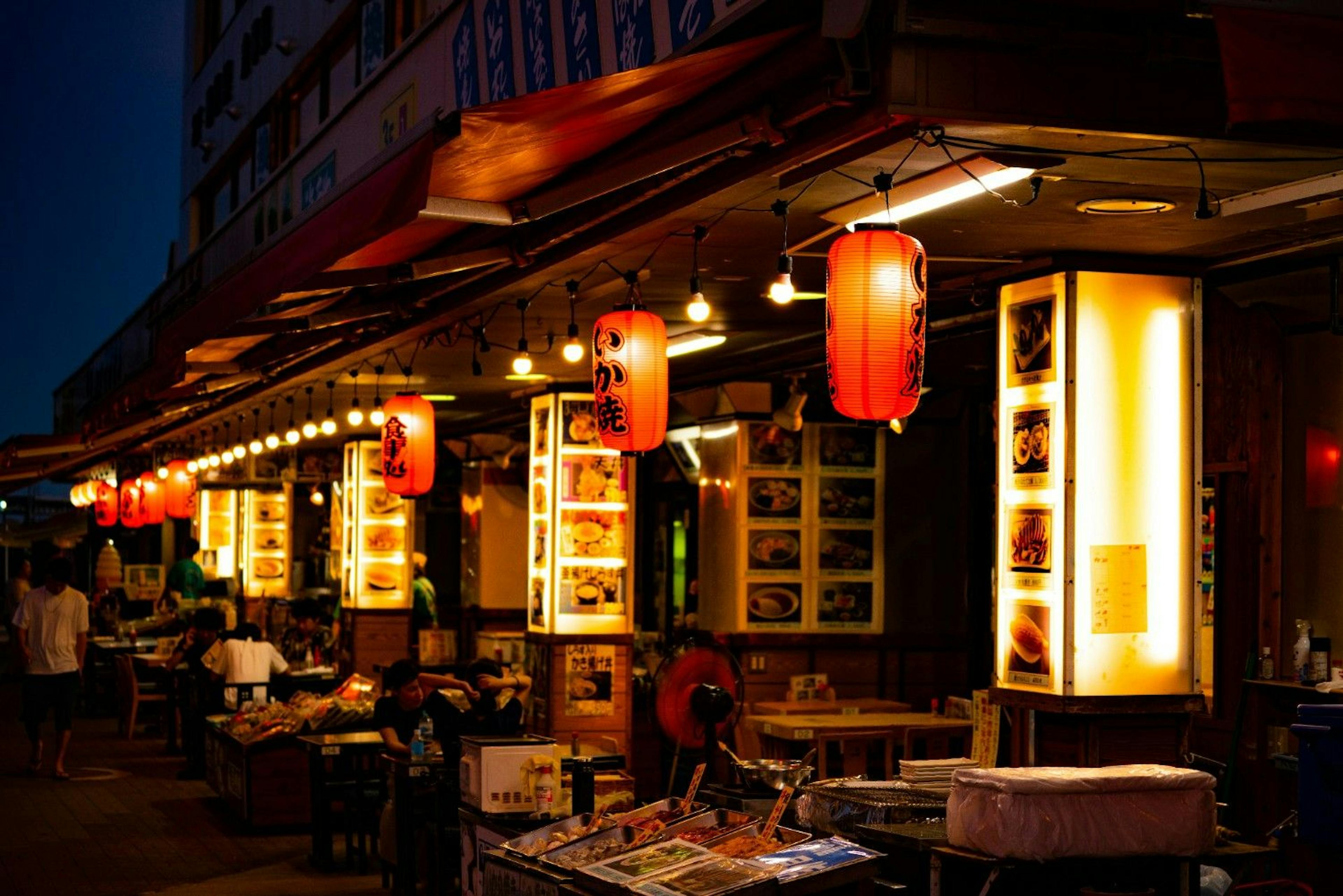 People eating at tables outside on a patio at a Japanese restaurant at night with lanterns and menus on column lights 