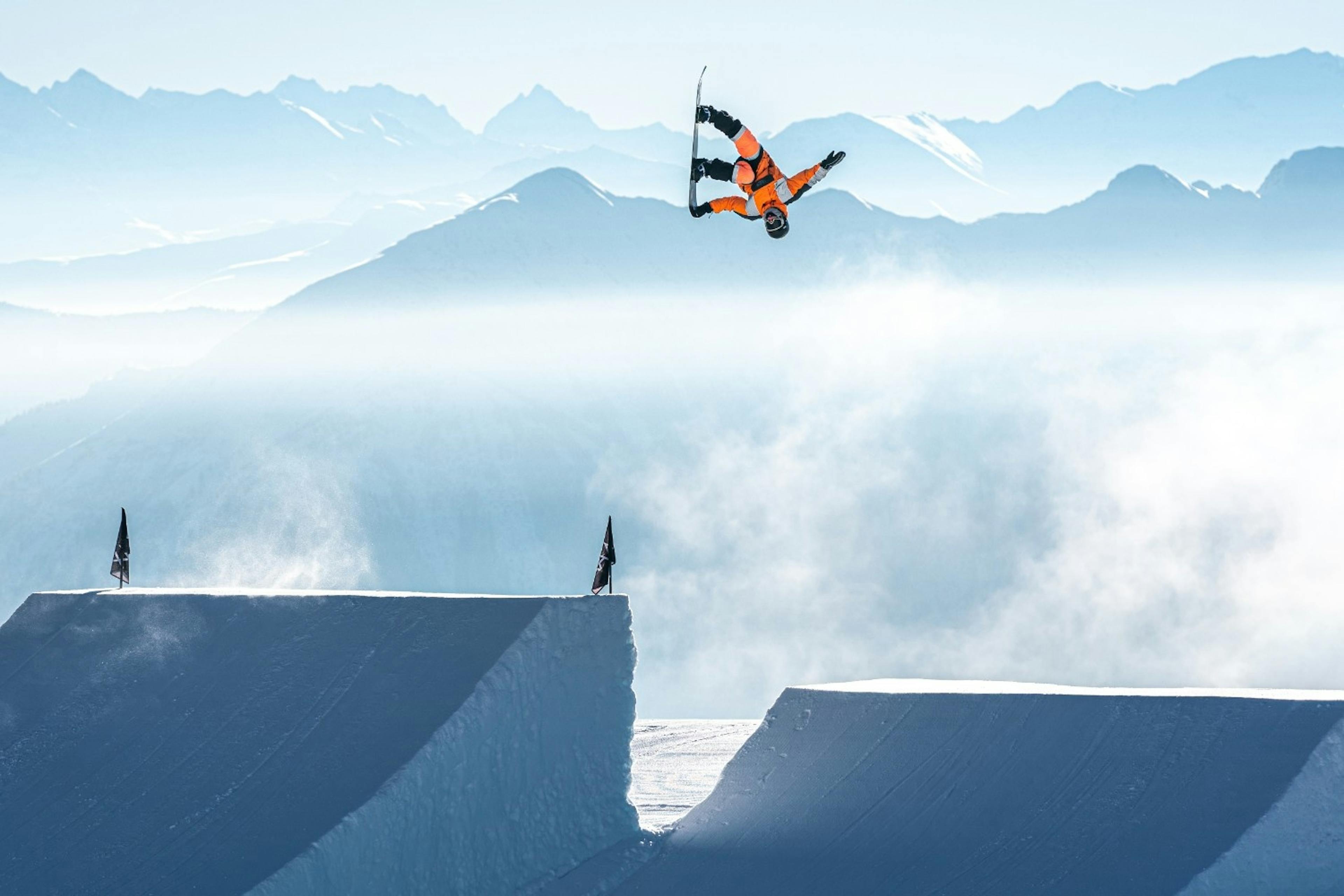 Snowboarder doing a backflip in the air in front of alpine peaks at LAAX Terrain Park