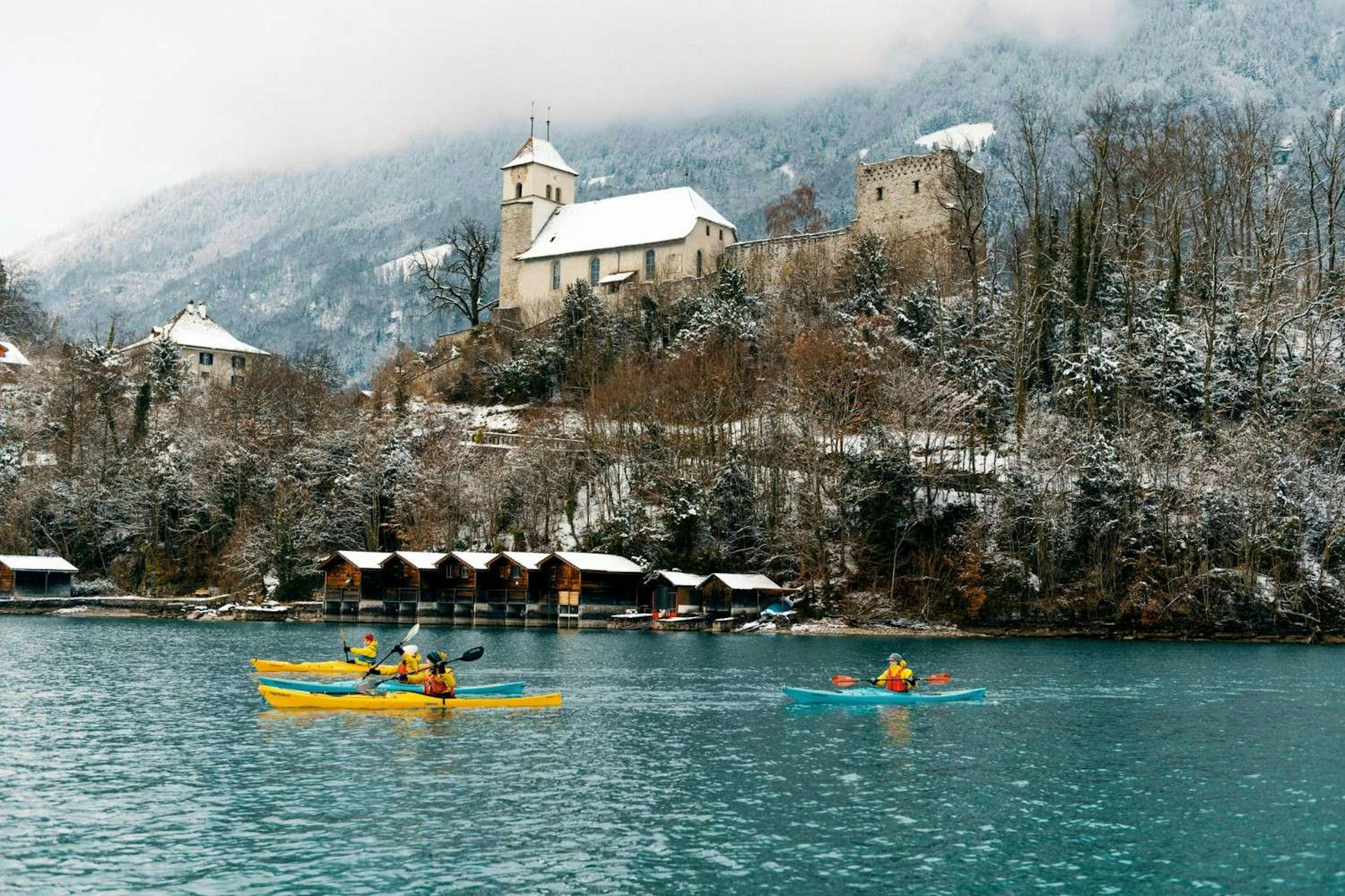 Three winter kayakers on the beautiful blue lake overlooking snowy castles, fog, hillsides, and trees 