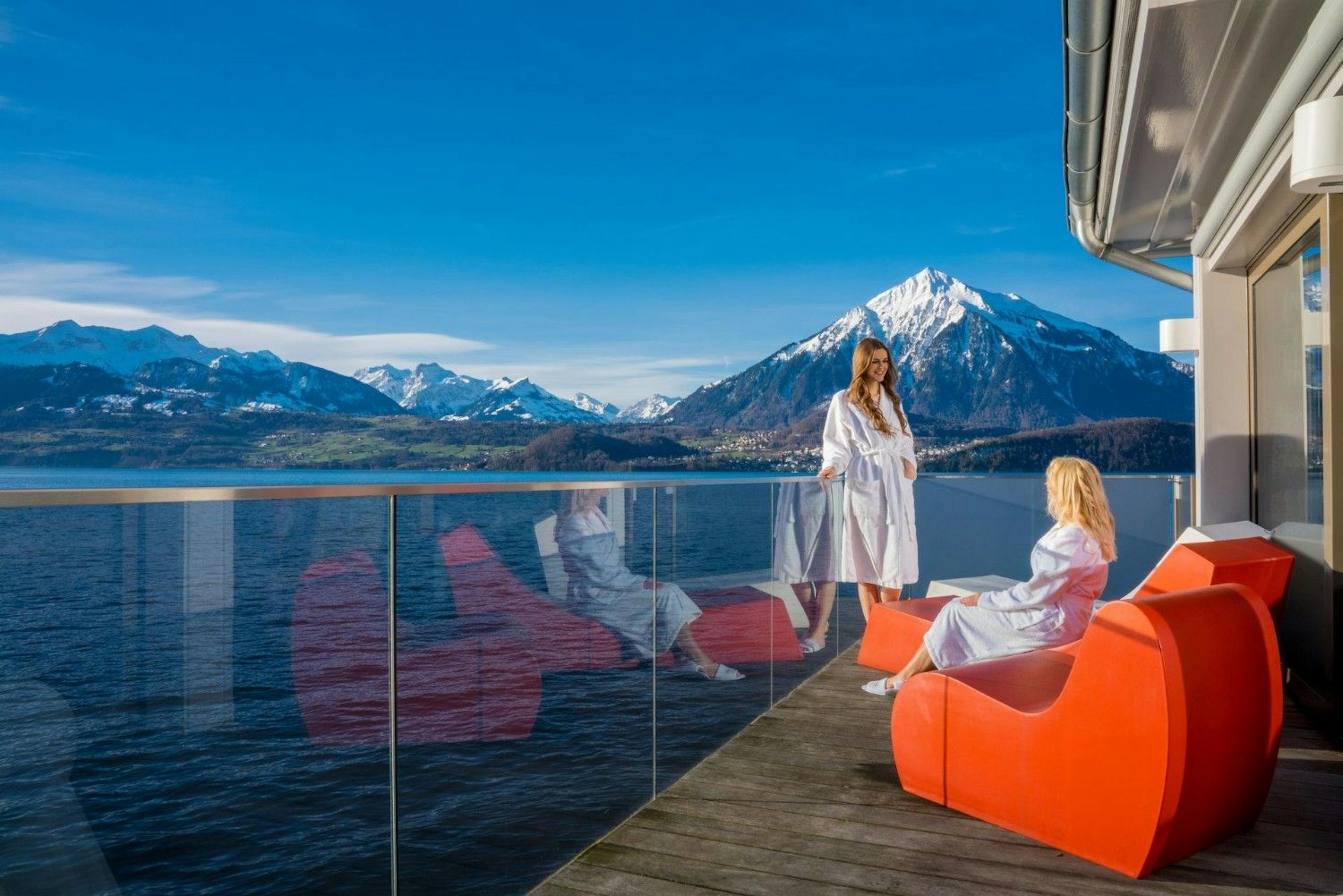 Two women enjoy a sunny patio in bathrobes overlooking the lake framed by snowy alpine mountains