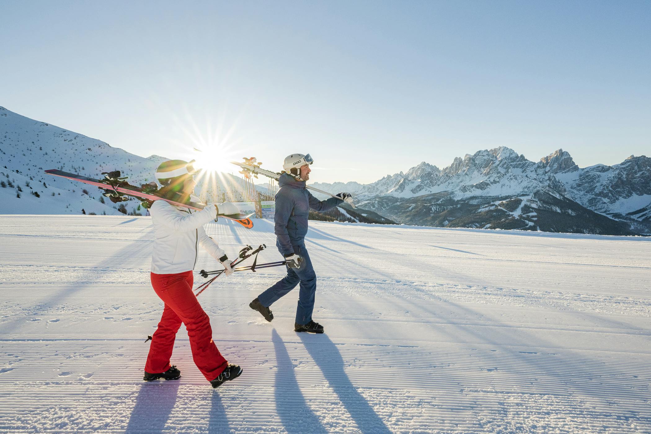 Male and female skier in colorful ski attire with ski sloped over their shoulder walking down the slopes at Three Peaks (3 Zinnen Dolomites) in Italy, Europe.