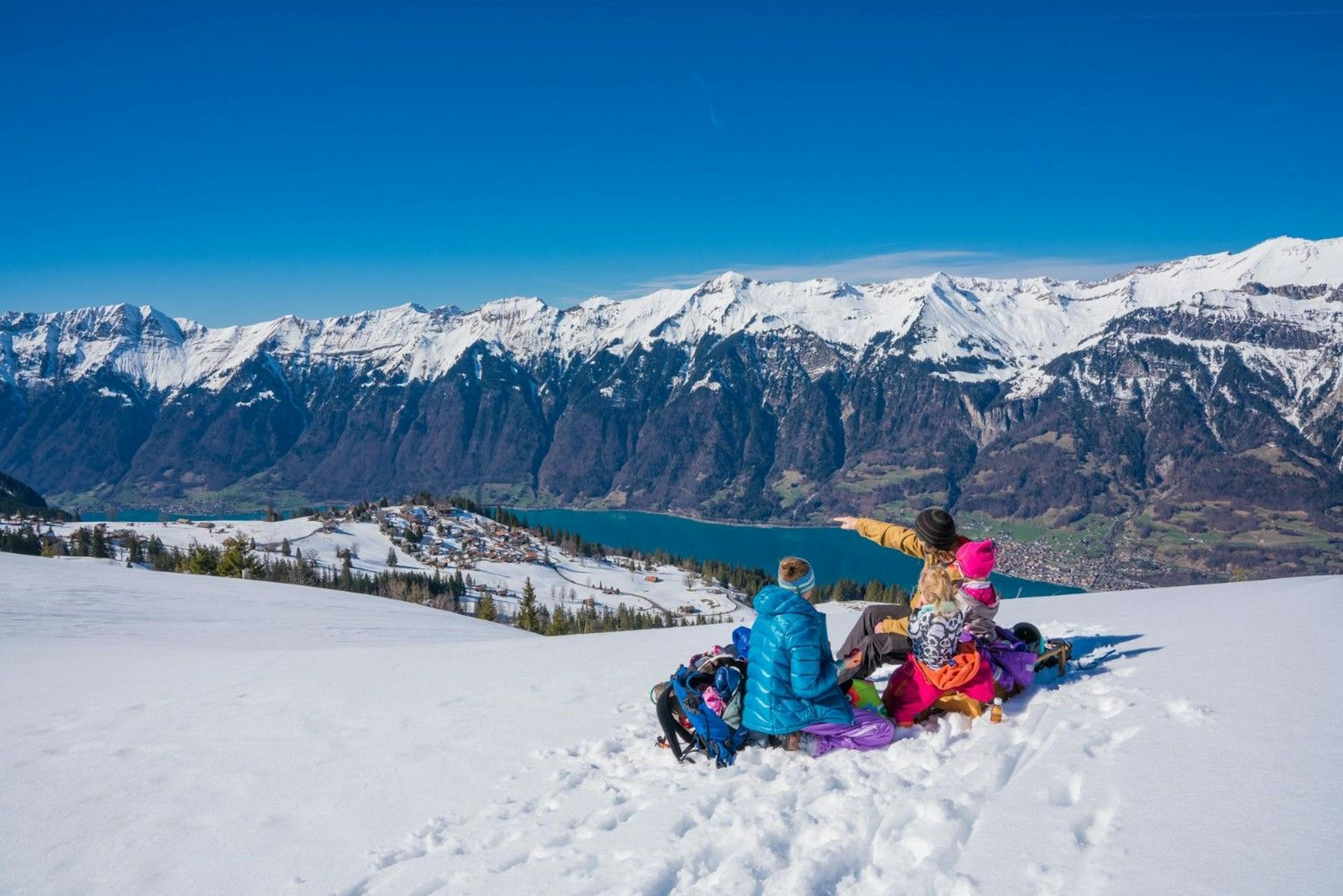 Family with small kids sitting on a snowy hill in snow gear overlooking the alpine mountains and Lake Brienz at Interlaken