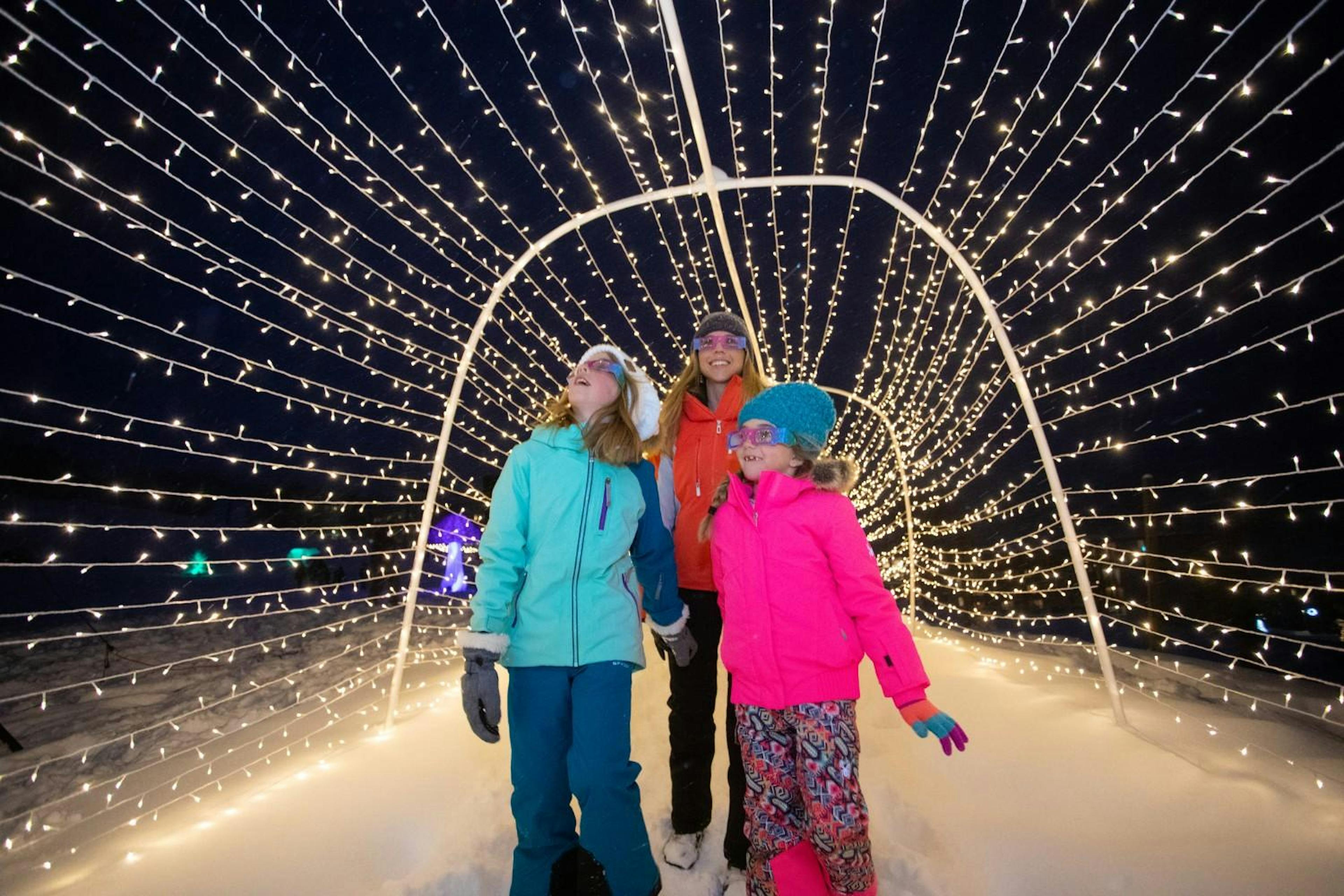 Mom and her two daughters walk under a tunnel of twinkly lights on a snowy night