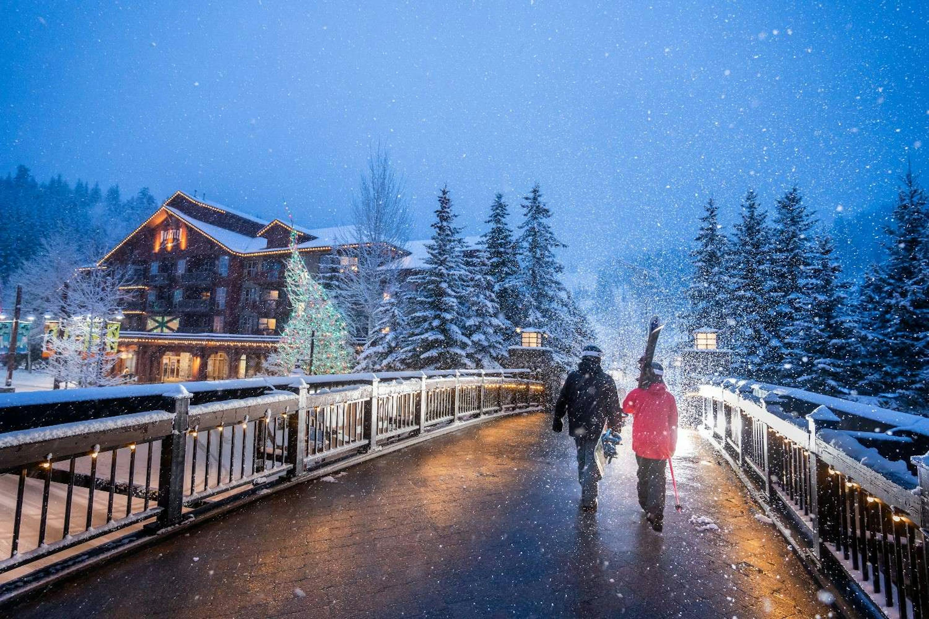 Two people in ski gear walk across a snowy bridge at twilight in Whistler Blackcomb Village