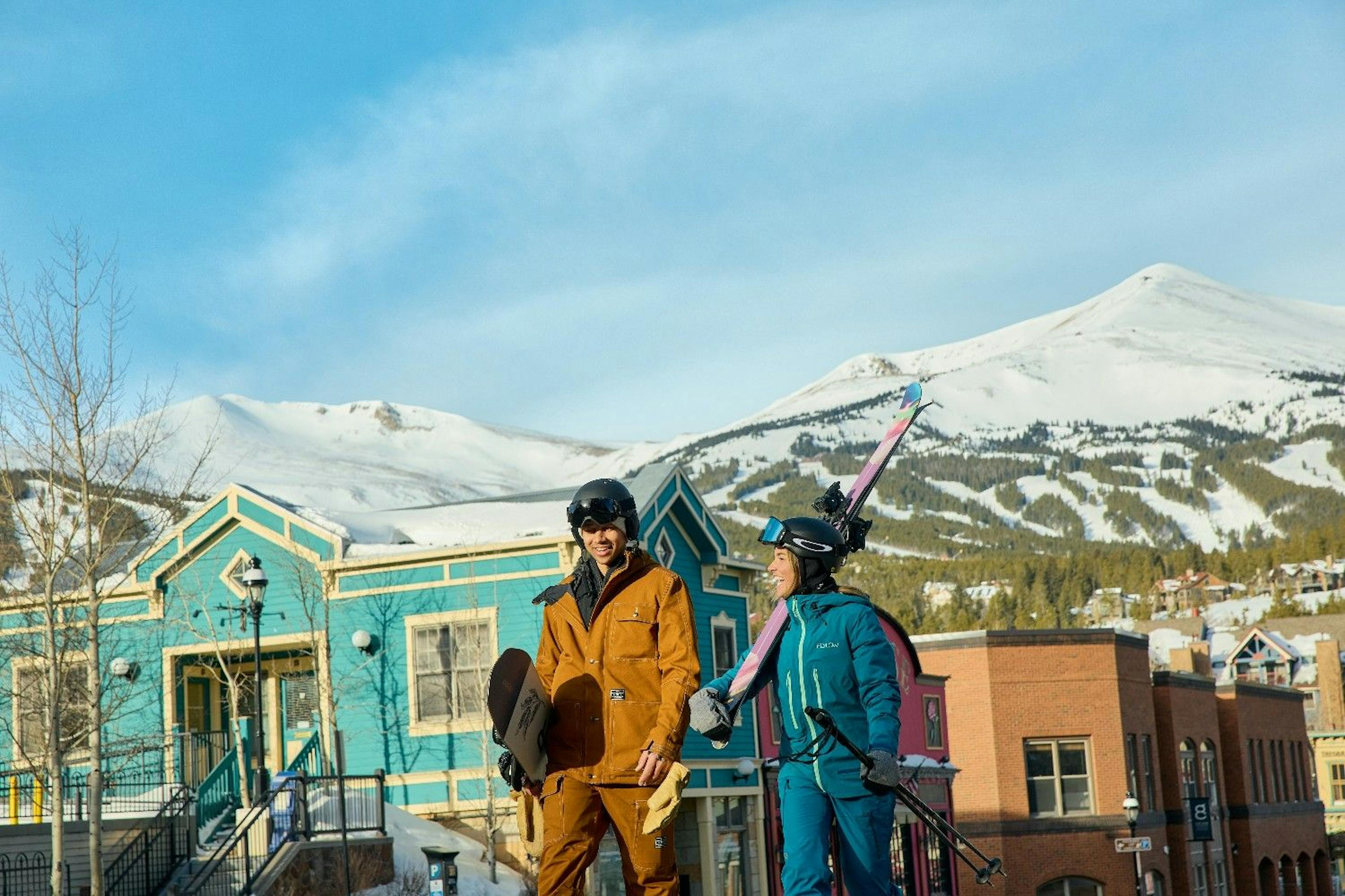 Two friends walk through the town of Breckenridge framed by the ski slopes with their ski and snowboard gear smiling on a sunny day