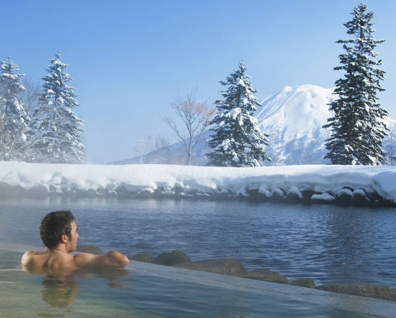 Man gazes at snow-covered Mount Yotei while soaking in an outdoor onsen in Niseko Village
