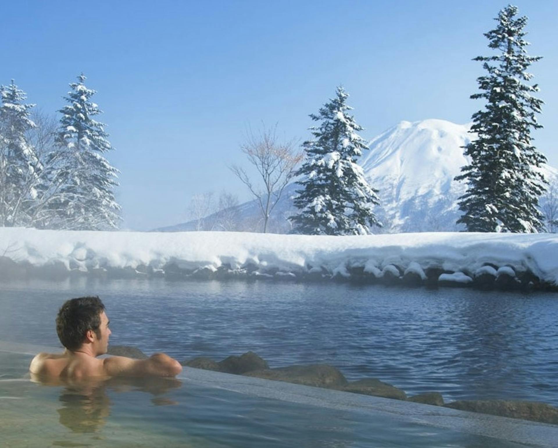 Man gazes at snow-covered Mount Yotei while soaking in an outdoor onsen in Niseko Village