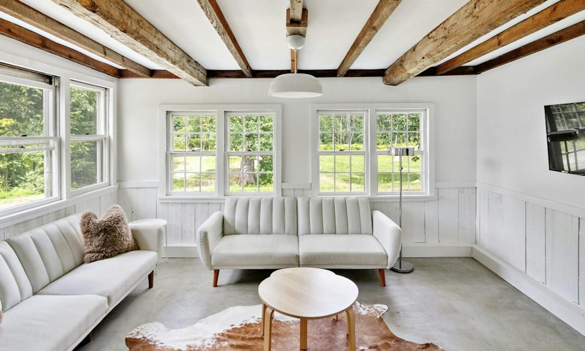 Inside a white cabin-like living room in a hotel room at Outbound Stowe with white modern couches and wrap-around windows with forest views
