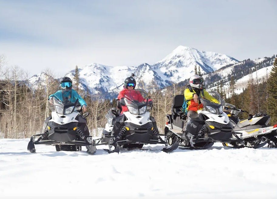 Three people in snow gear on snowmobiles with snowy alpine mountains behind them