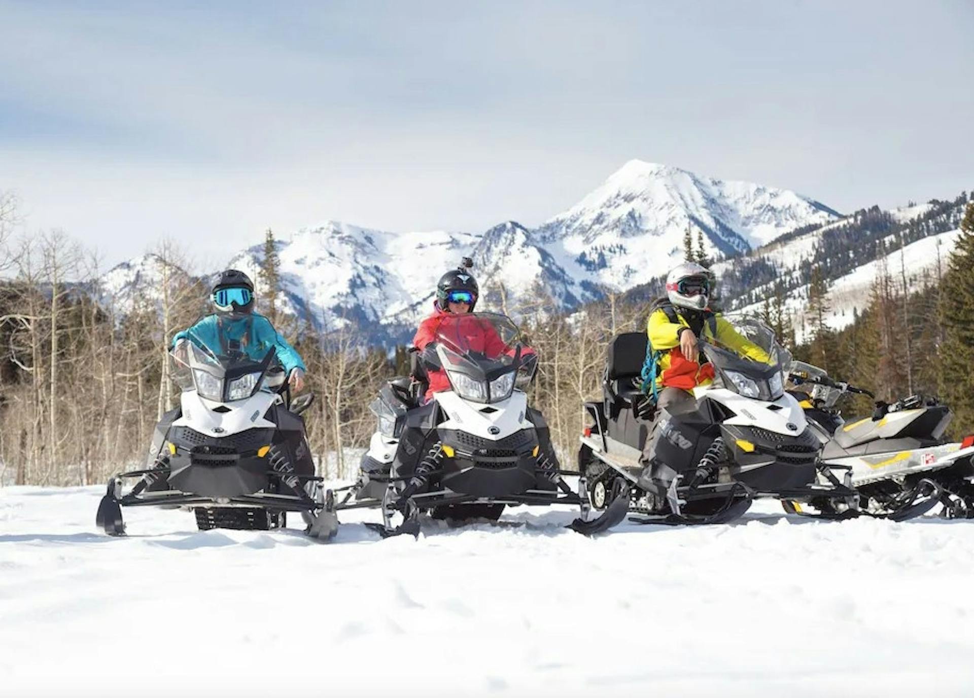 Three people in snow gear on snowmobiles with snowy alpine mountains behind them