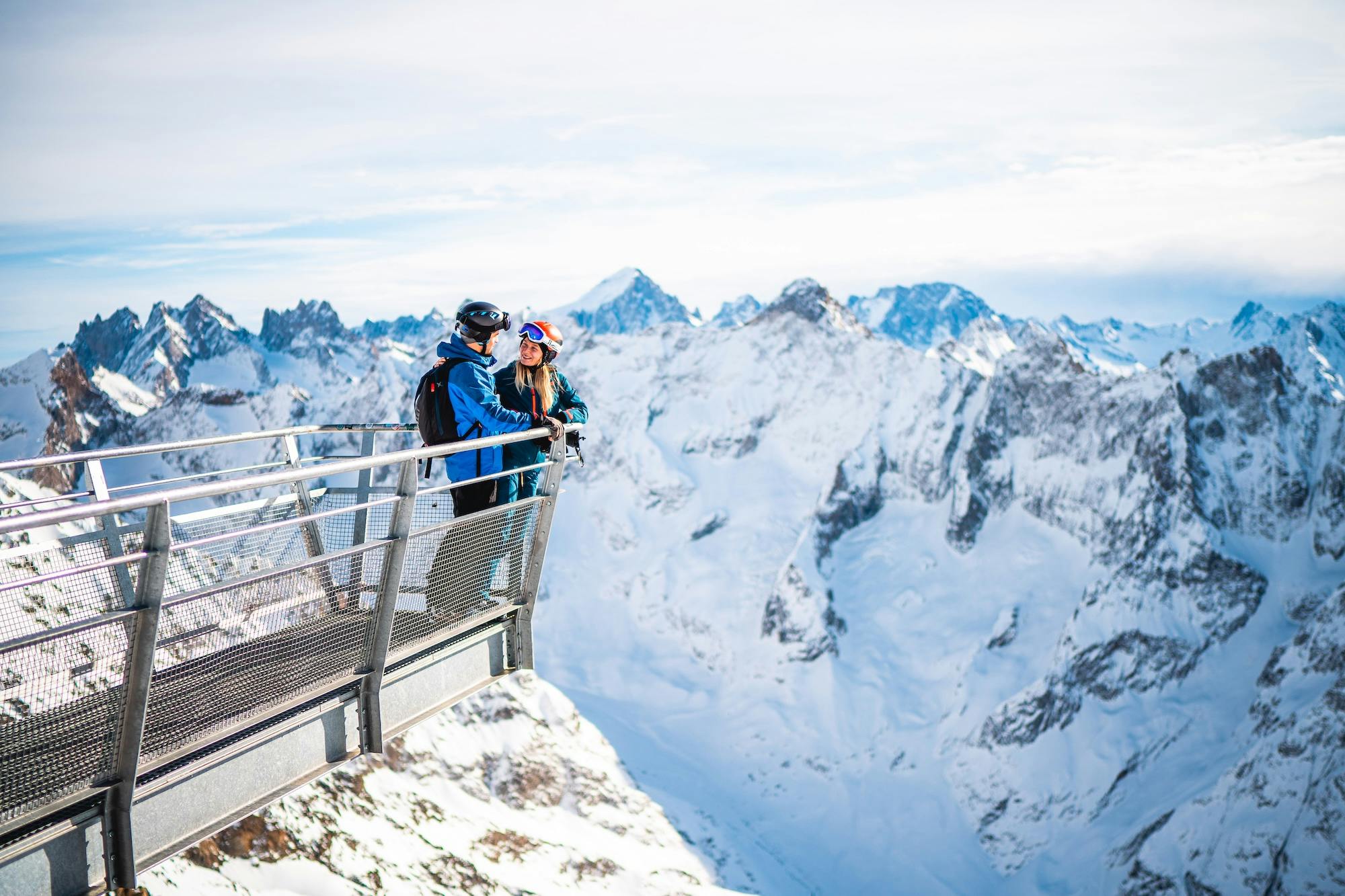 Two people stand on a viewing platform overlooking snowy mountain peaks