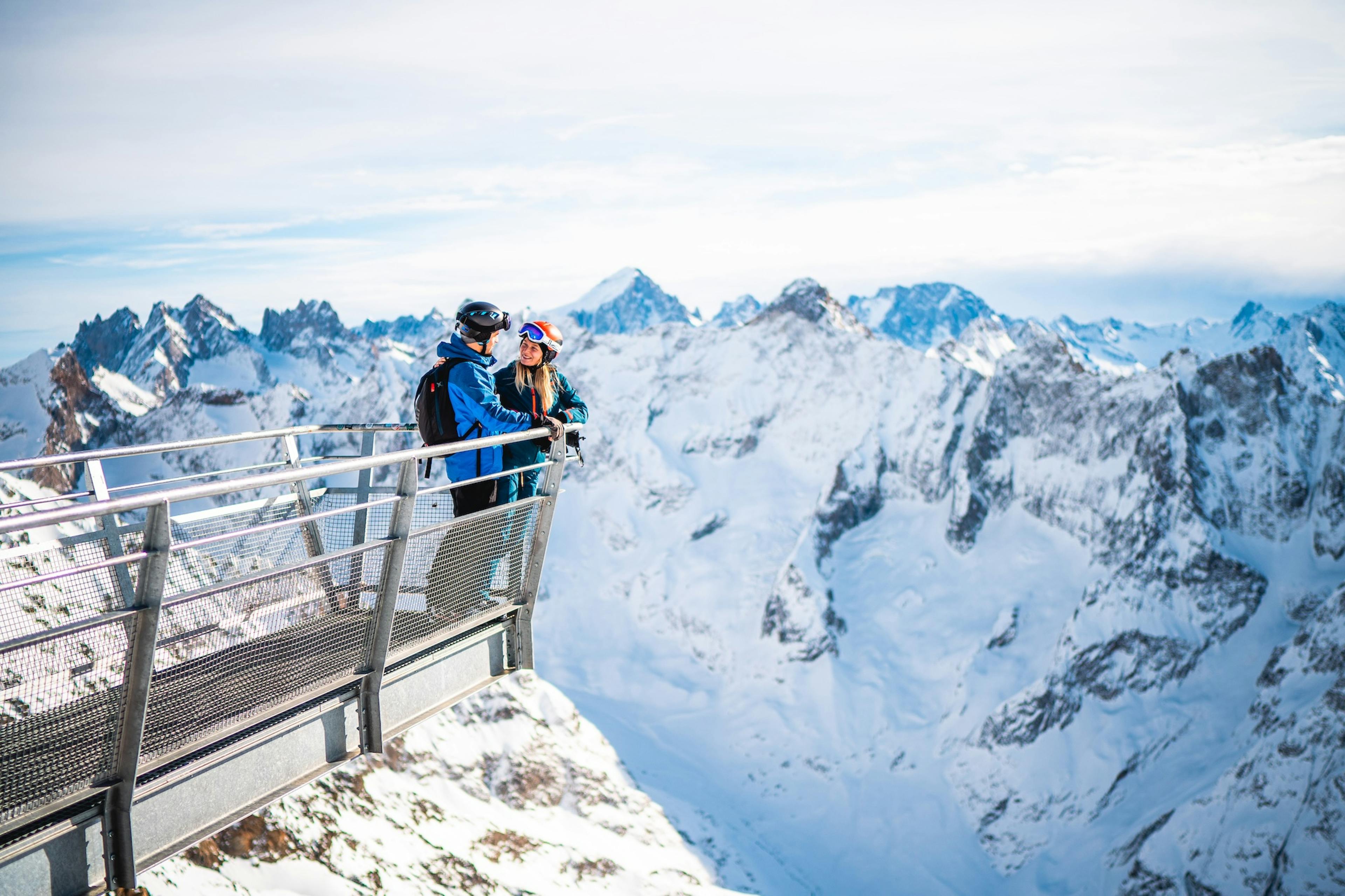 Two people stand on a viewing platform overlooking snowy mountain peaks