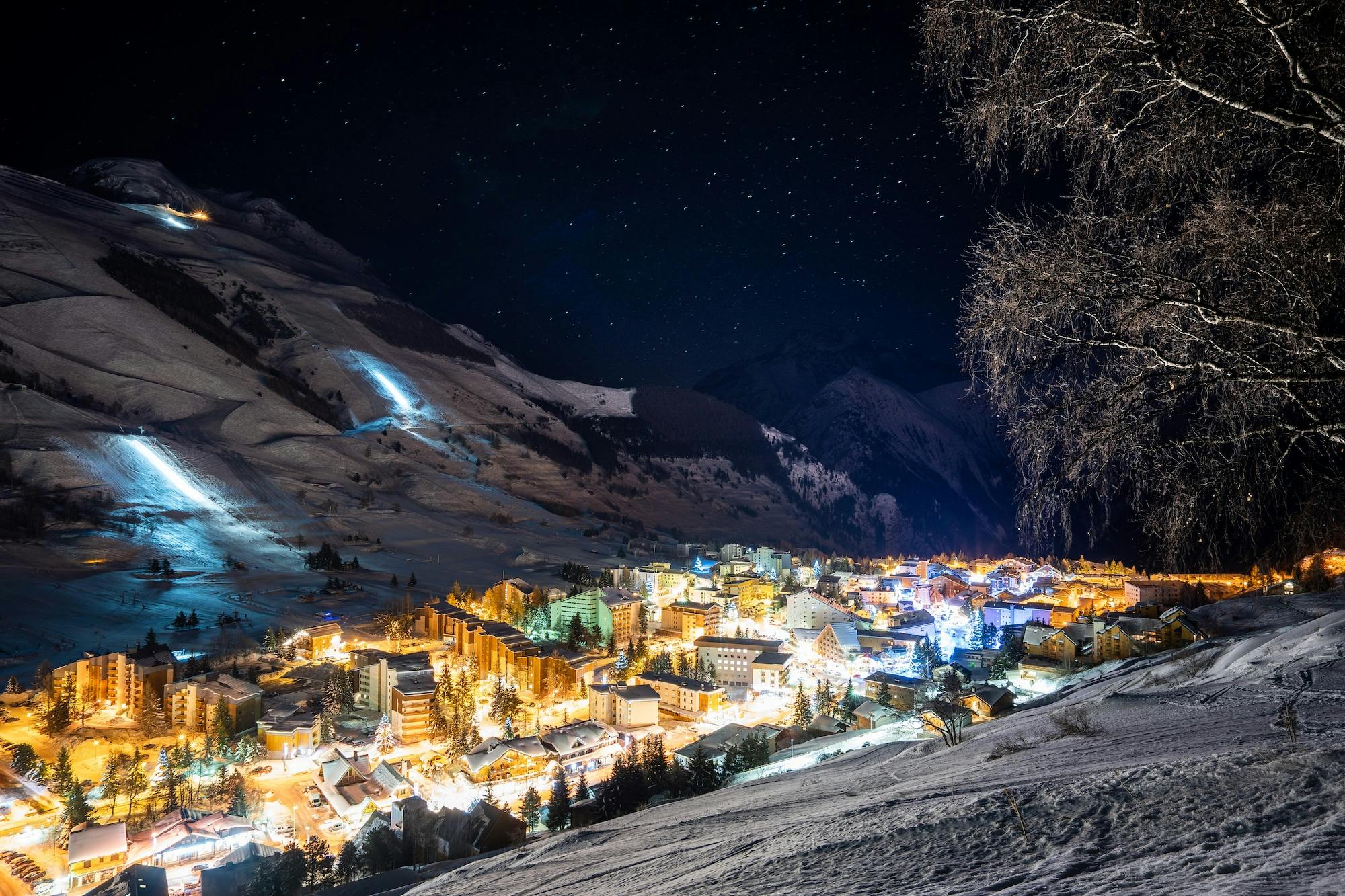 A snow-covered mountain town at night, illuminated by streetlights and building lights, with ski slopes visible on the hillside.