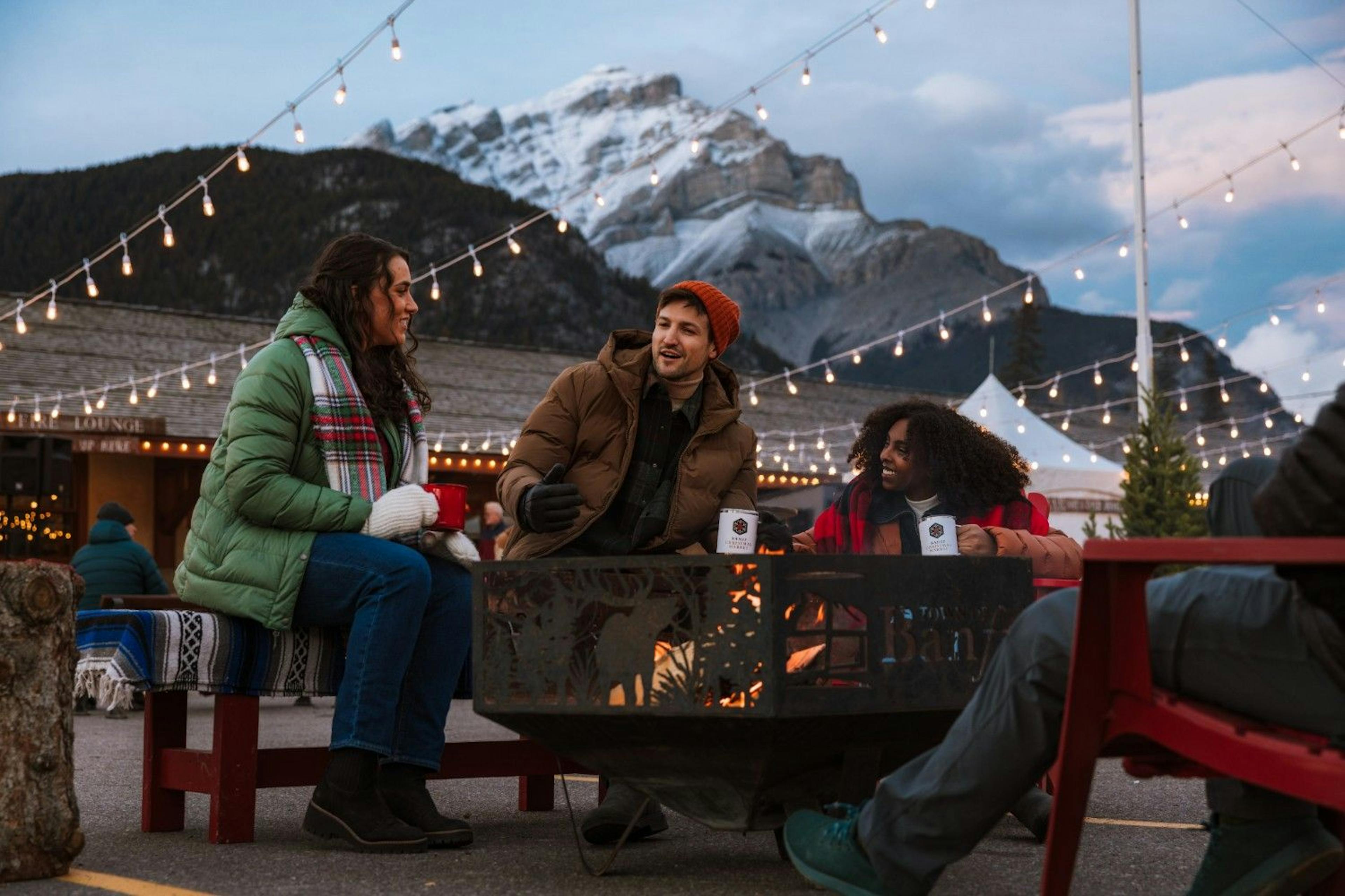 Three friends wearing winter clothes sit outside around a firepit under string lights framed by the Canadian Rockies