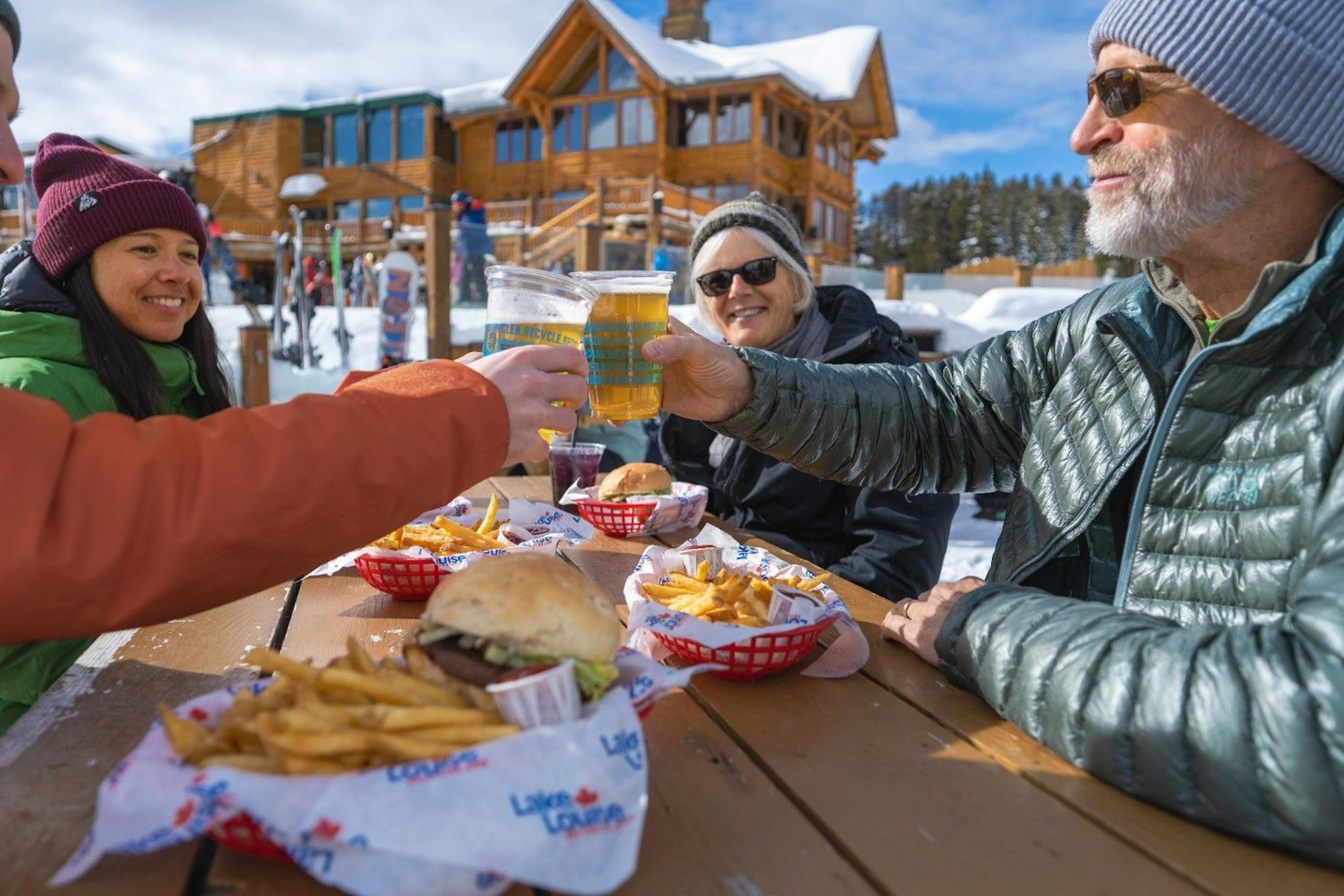Group of four cheers their beers over burgers sitting on a sunny patio of a ski lodge at Lake Louise