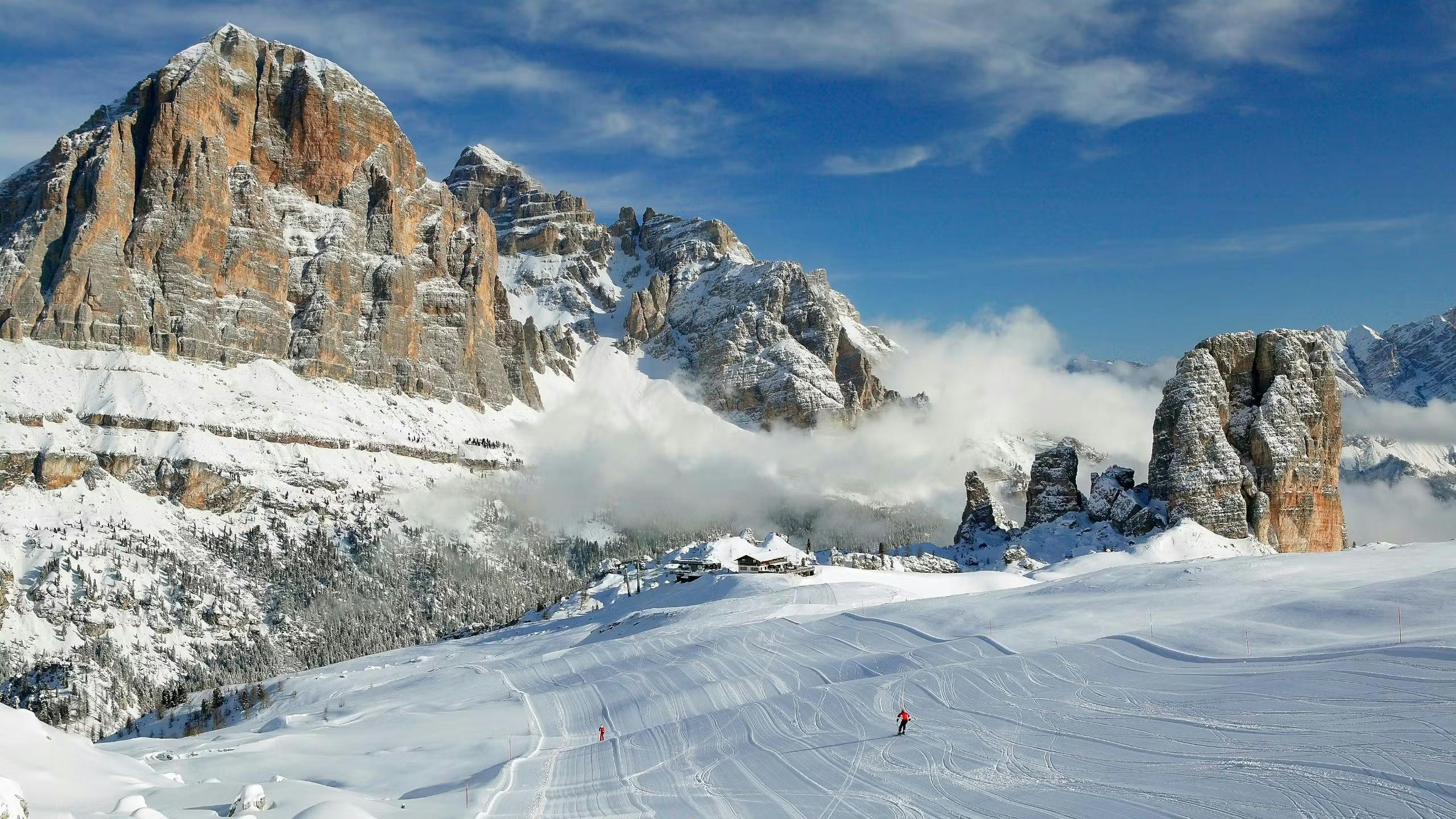 The sun sets over the snowy mountains and slopes of Cortina d'Ampezzo as two people ski down the run