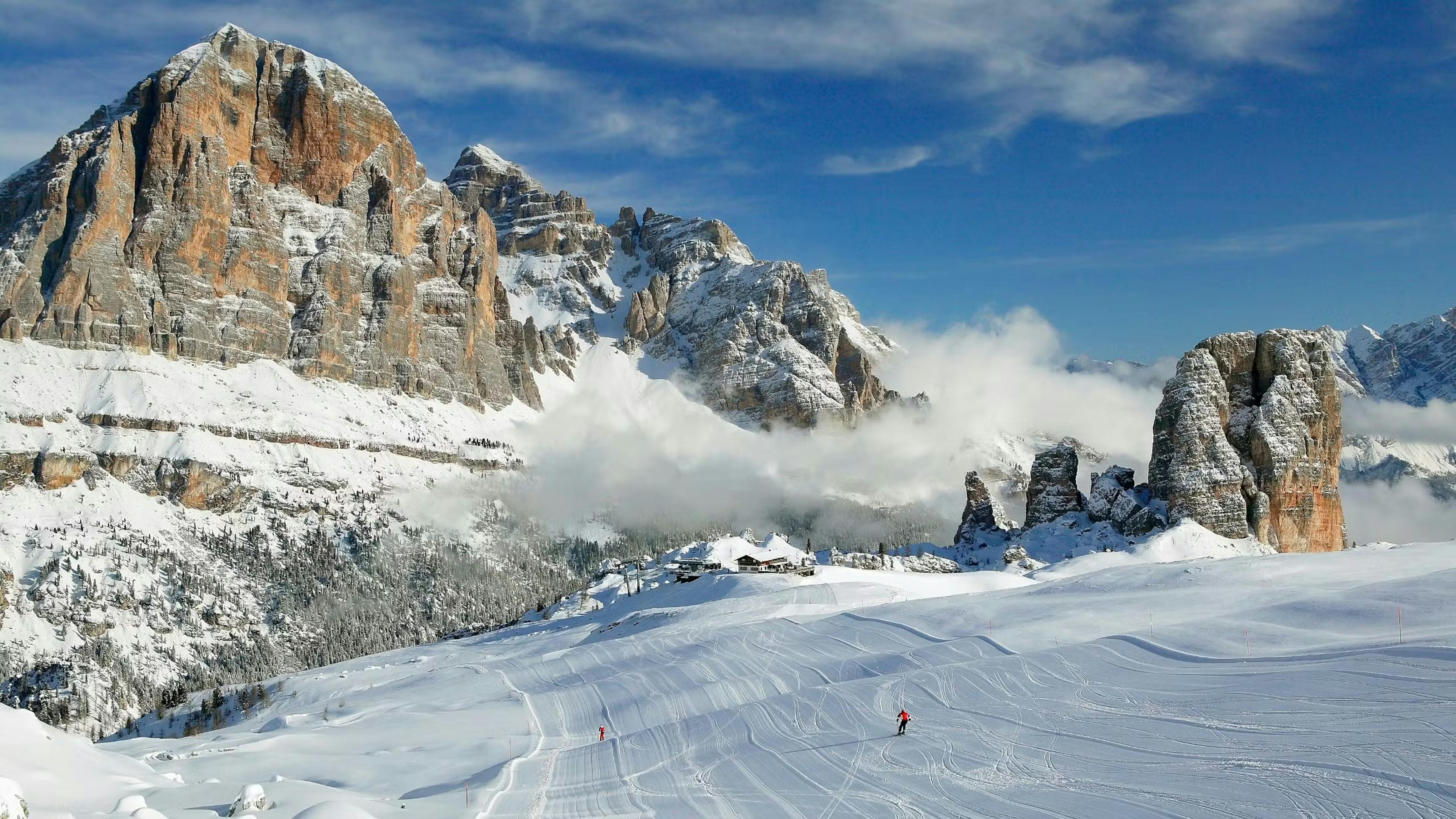 The sun sets over the snowy mountains and slopes of Cortina d'Ampezzo as two people ski down the run