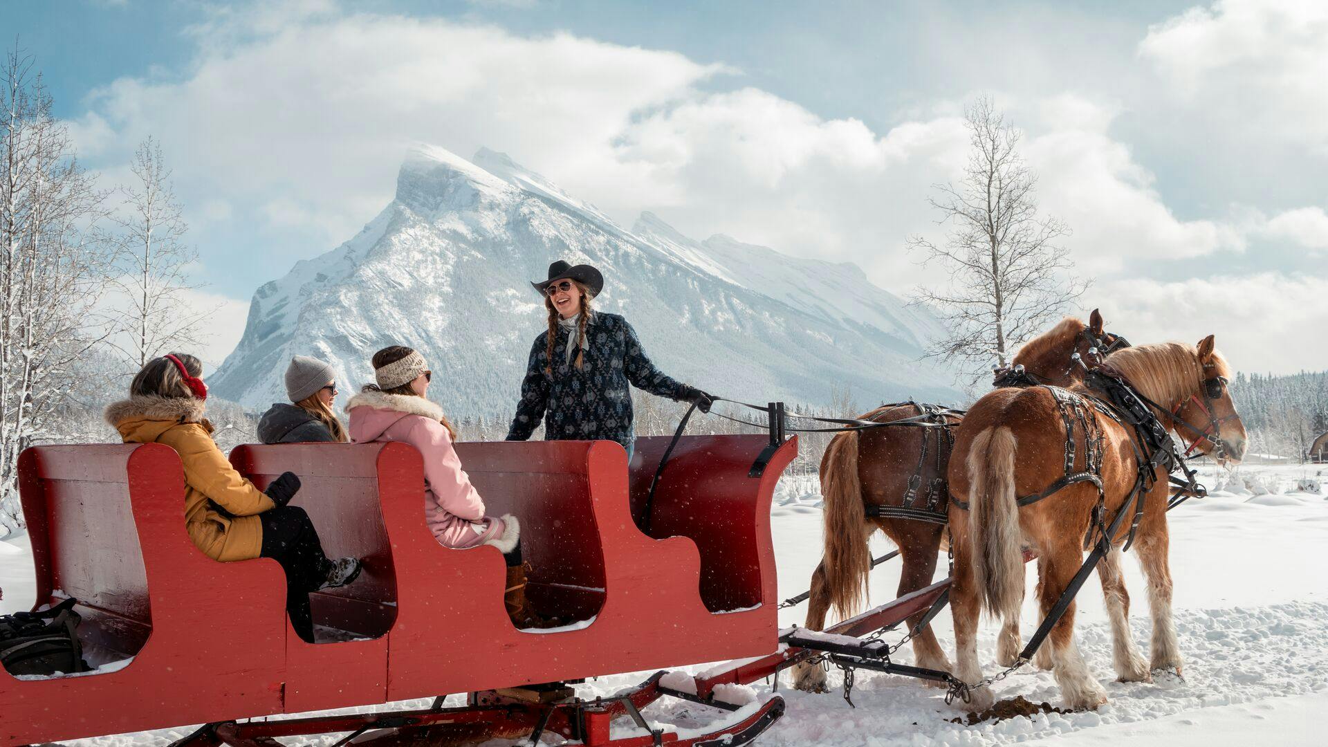 A woman wearing a cowgirl hat guides two horses pulling a sleigh of people in the snow, framed by snowy alpine mountains