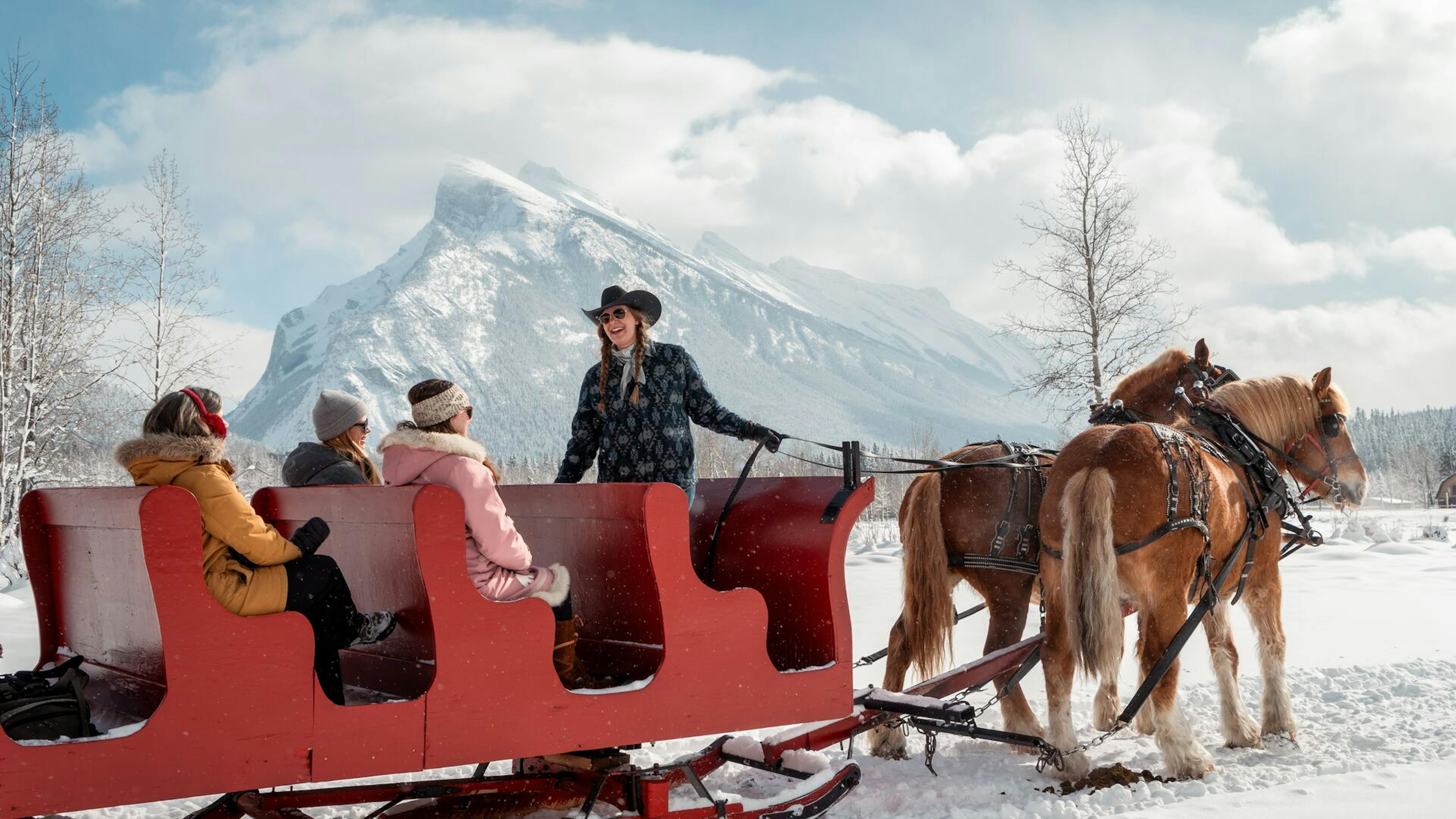 A woman wearing a cowgirl hat guides two horses pulling a sleigh of people in the snow, framed by snowy alpine mountains