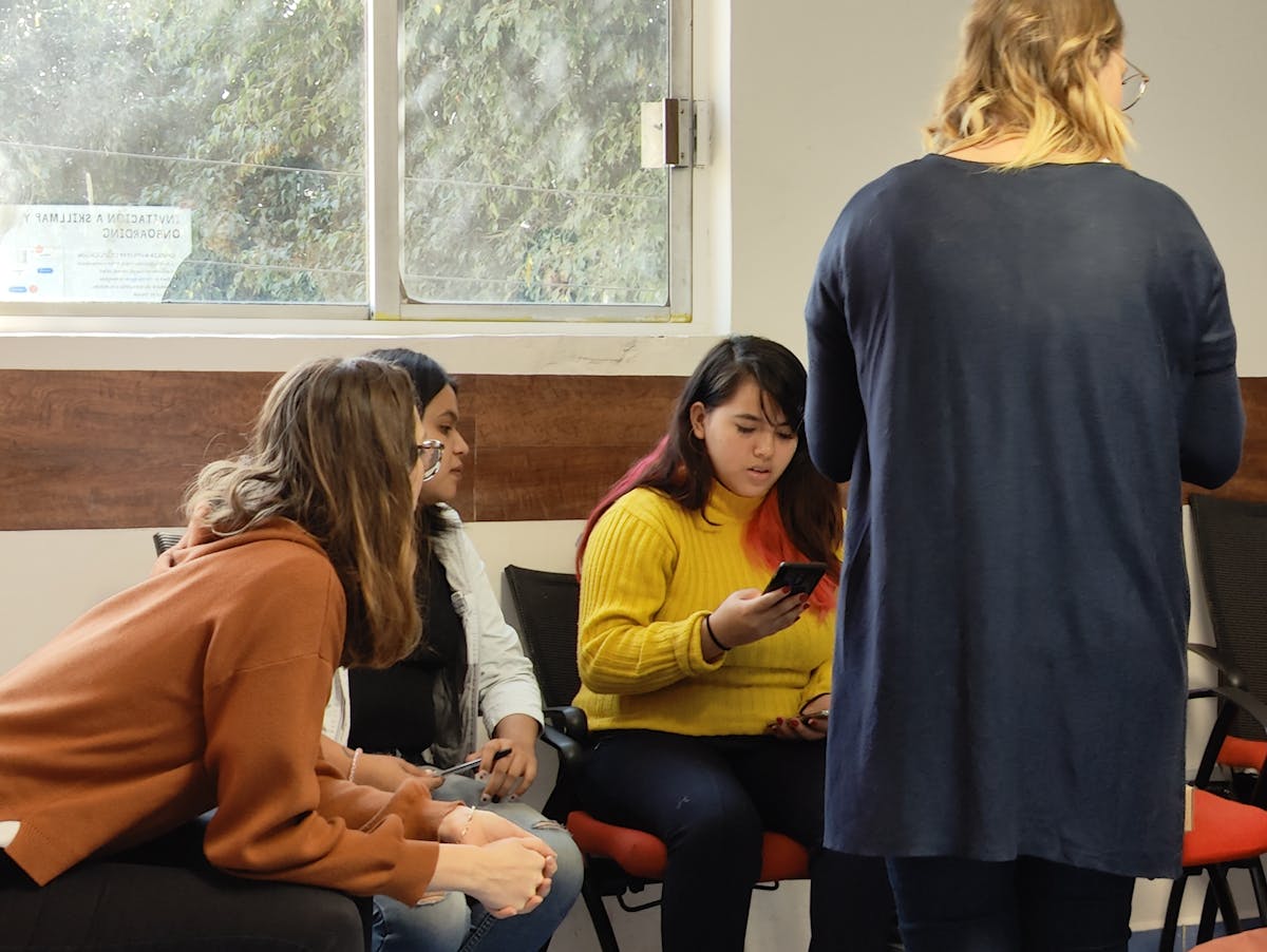 Four women, three sitting and one standing, discussing the app while one of them is using it on the phone