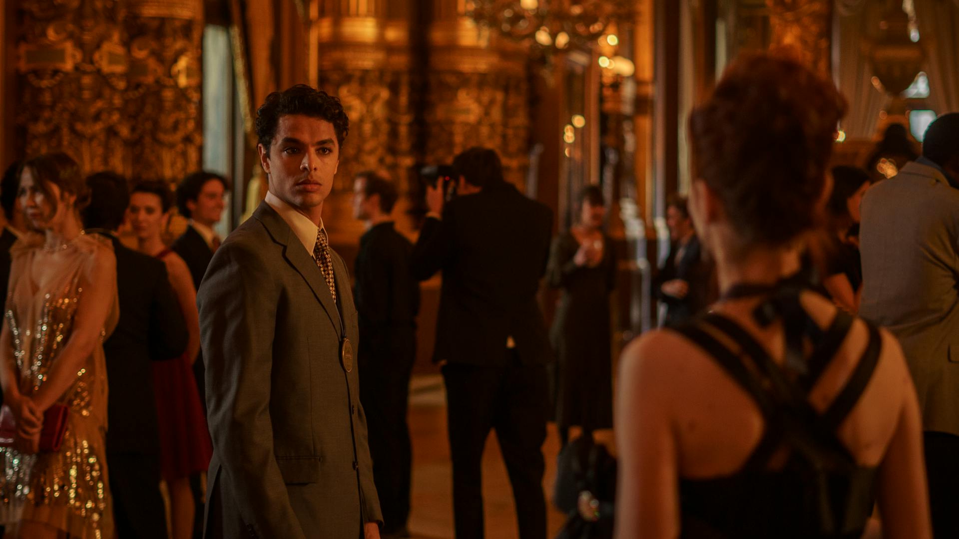 Thiago Soares (Matheus Abreu) está parado de terno em frente à tela, em meio a uma festa com algumas pessoas. Ele encara uma mulher que está de costas para a tela.