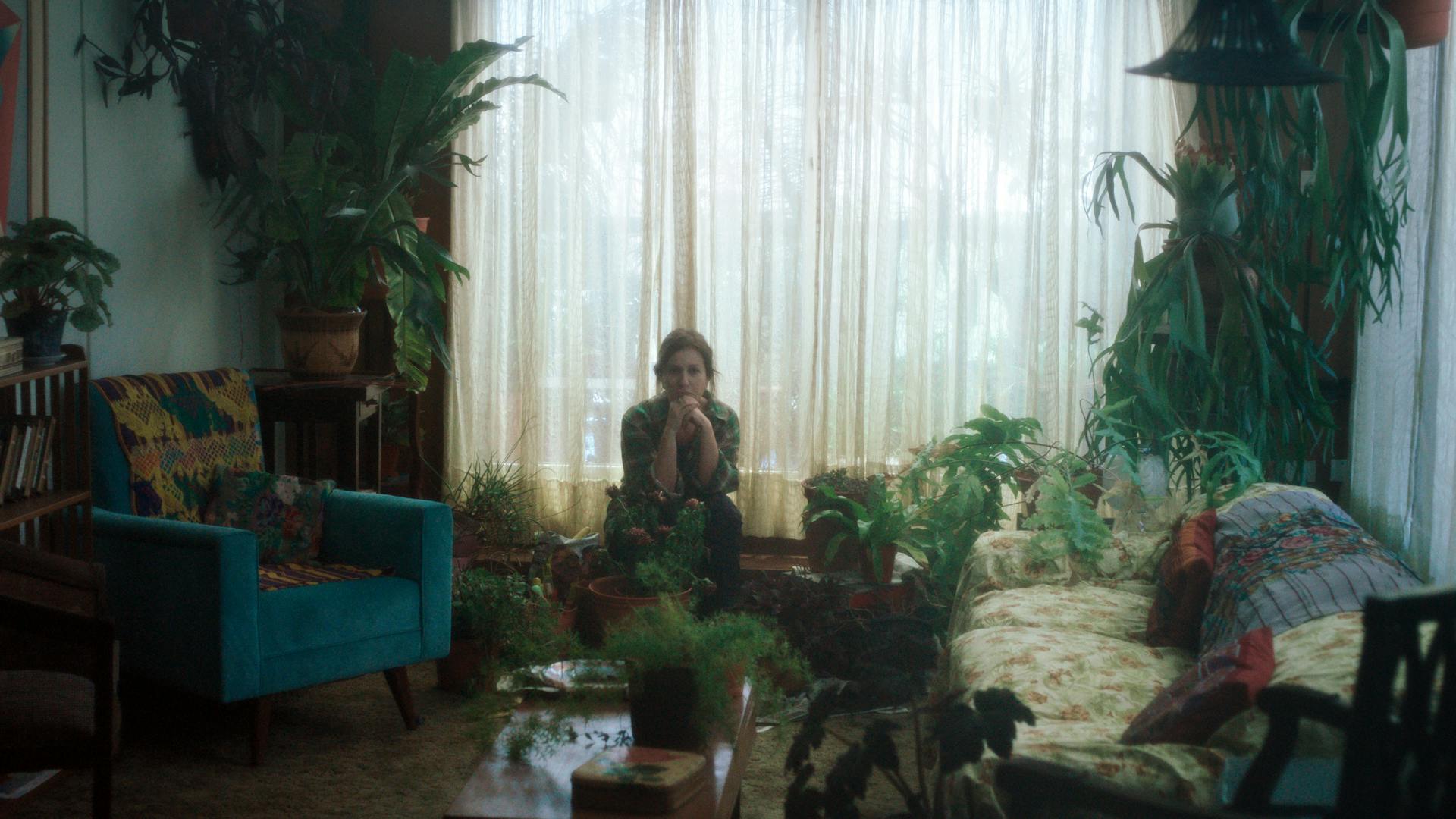 Uma mulher está sentada em uma sala de estar, com poltronas, sofá e muitas plantas ao redor.