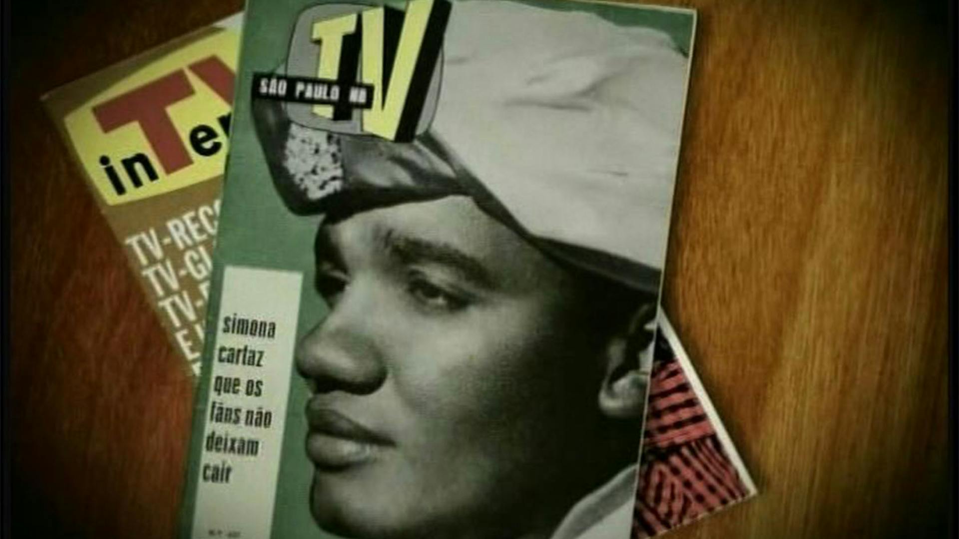 Diversas revistas sobre uma mesa de madeira, com a capa em destaque mostrando uma foto preto e branco, com fundo verde, com o rosto de Simonal de lado olhando para esquerda