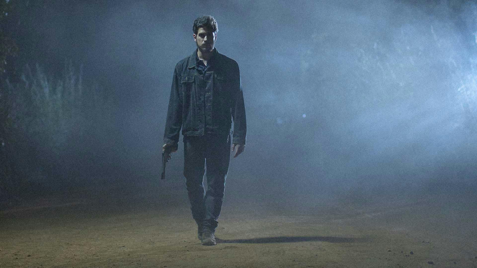 Marco Pigossi está em pé e segura uma pistola na mão em meio à névoa da noite.