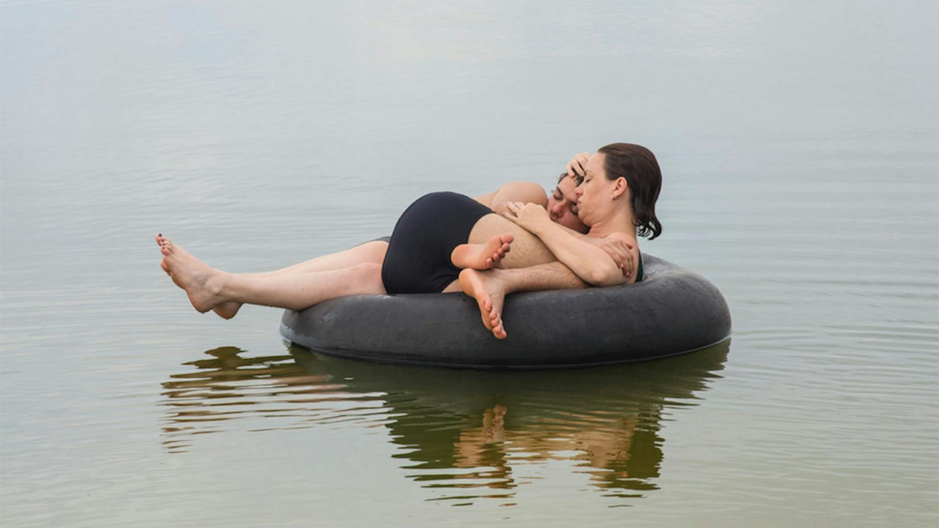 Uma mãe segura seu filho adolescente nos braços. Ambos estão em uma boia redonda dentro do mar.