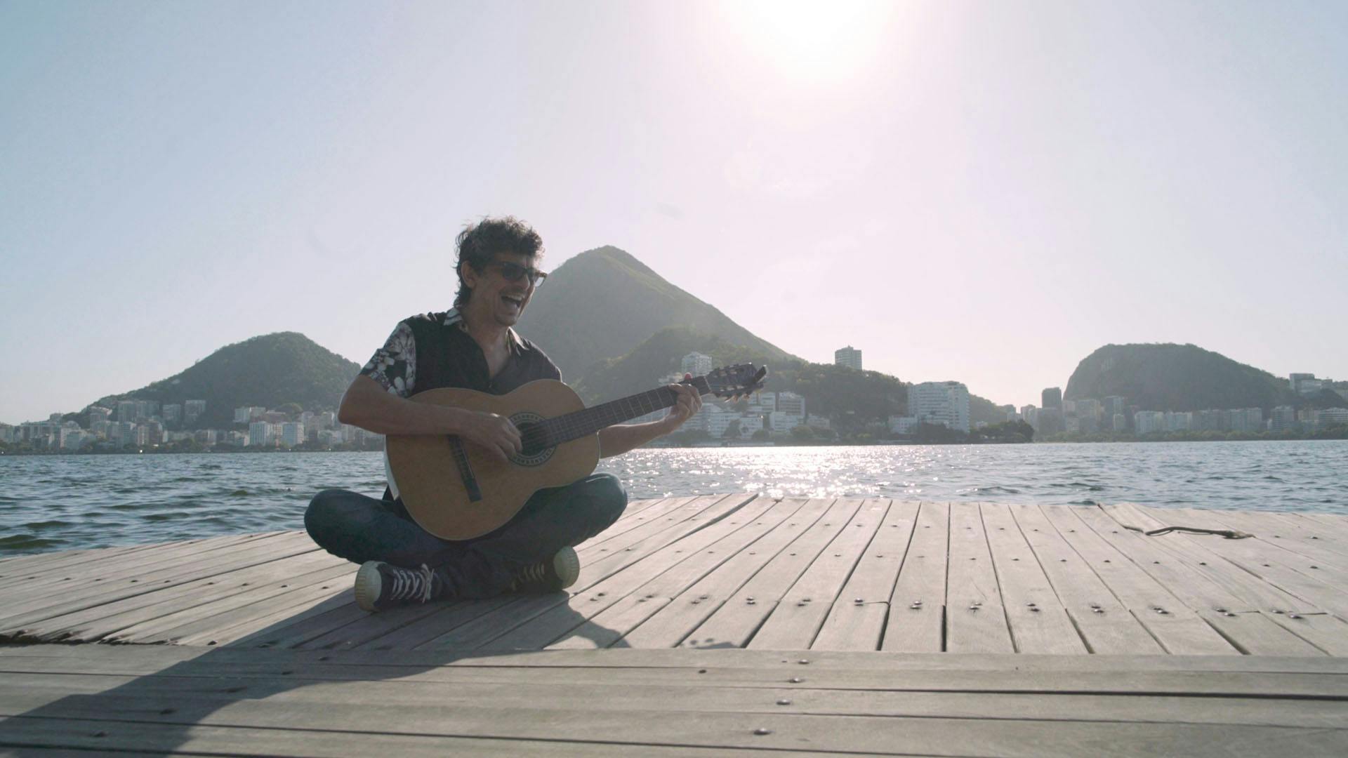 Pedro Luis está sentado e tocando violão em um deck de frente para a Baía do Rio de Janeiro com o Pão de Açucar ao fundo.