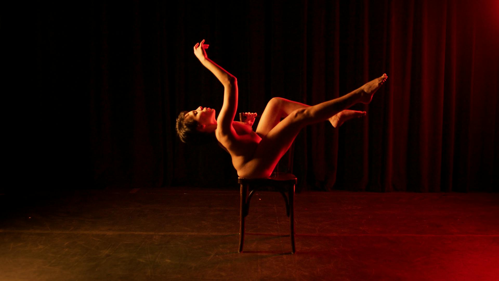 Uma mulher está nua, posando em cima de uma cadeira, com um dos braços erguidos para cima