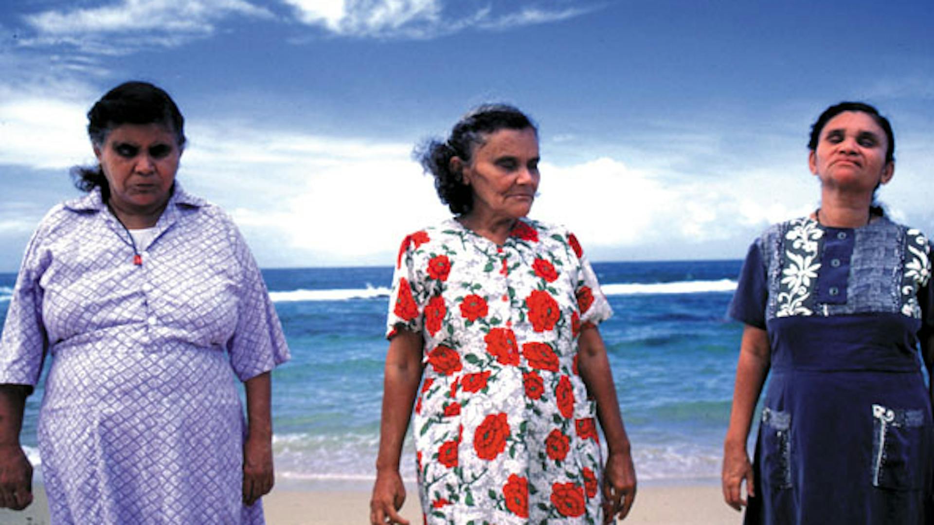 Três senhoras cegas estão de frente para a tela. Ao horizonte, o céu e mar azul.