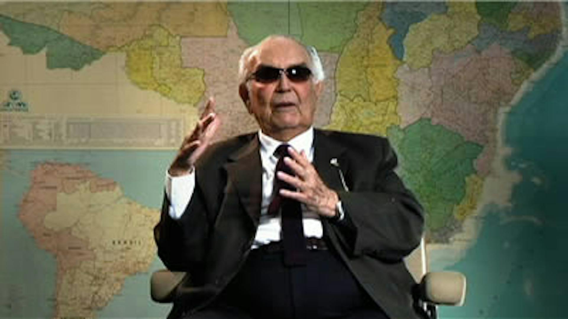 Eliezer Batista está de terno e óculos escuros, sentado em uma cadeira. Ao fundo, um mapa do mundo.