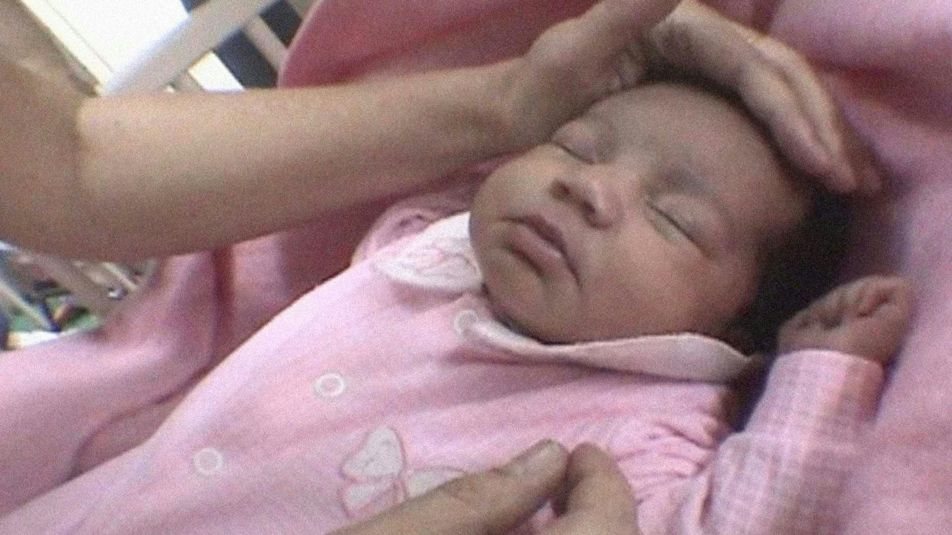 Um bebê recém-nascido com roupas rosas está dormindo. Uma mão está passando acariciando sua cabeça.