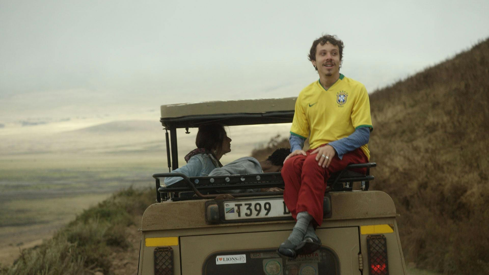 Homem com a camisa da seleção brasileira sentado na caçamba de um jipe, com horizonte de montanhas ao fundo.